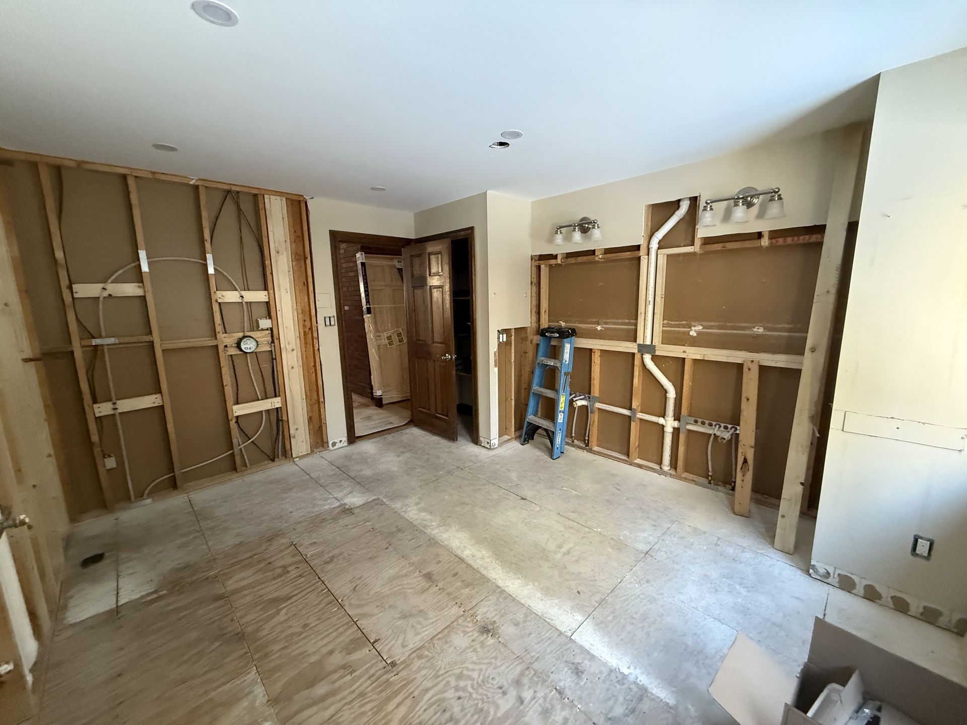 Kitchen undergoing renovation; walls stripped to studs, plywood floor visible, ladder present.