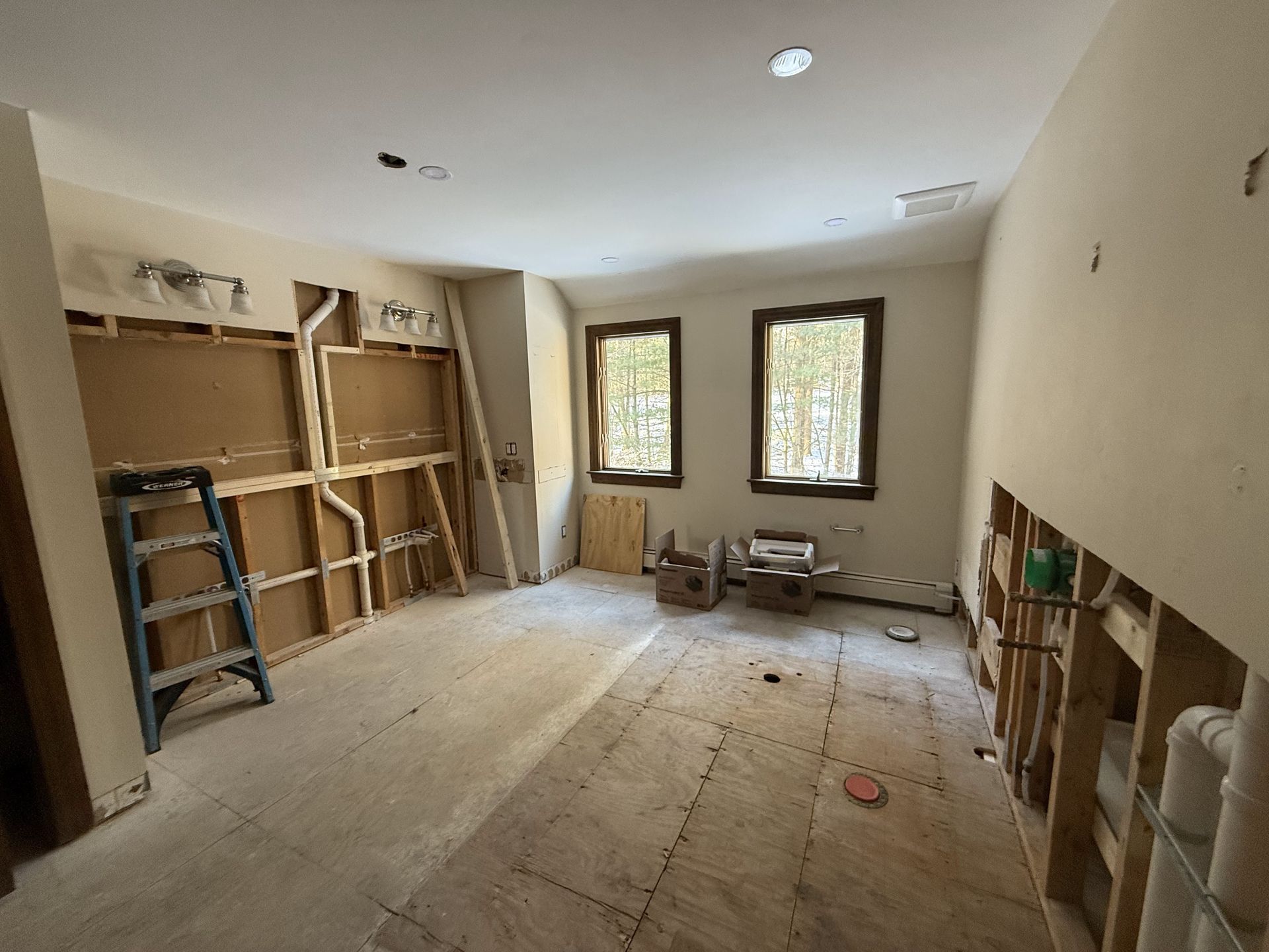 Room under construction; exposed wall studs, two windows, ladder, and tools scattered on the floor.