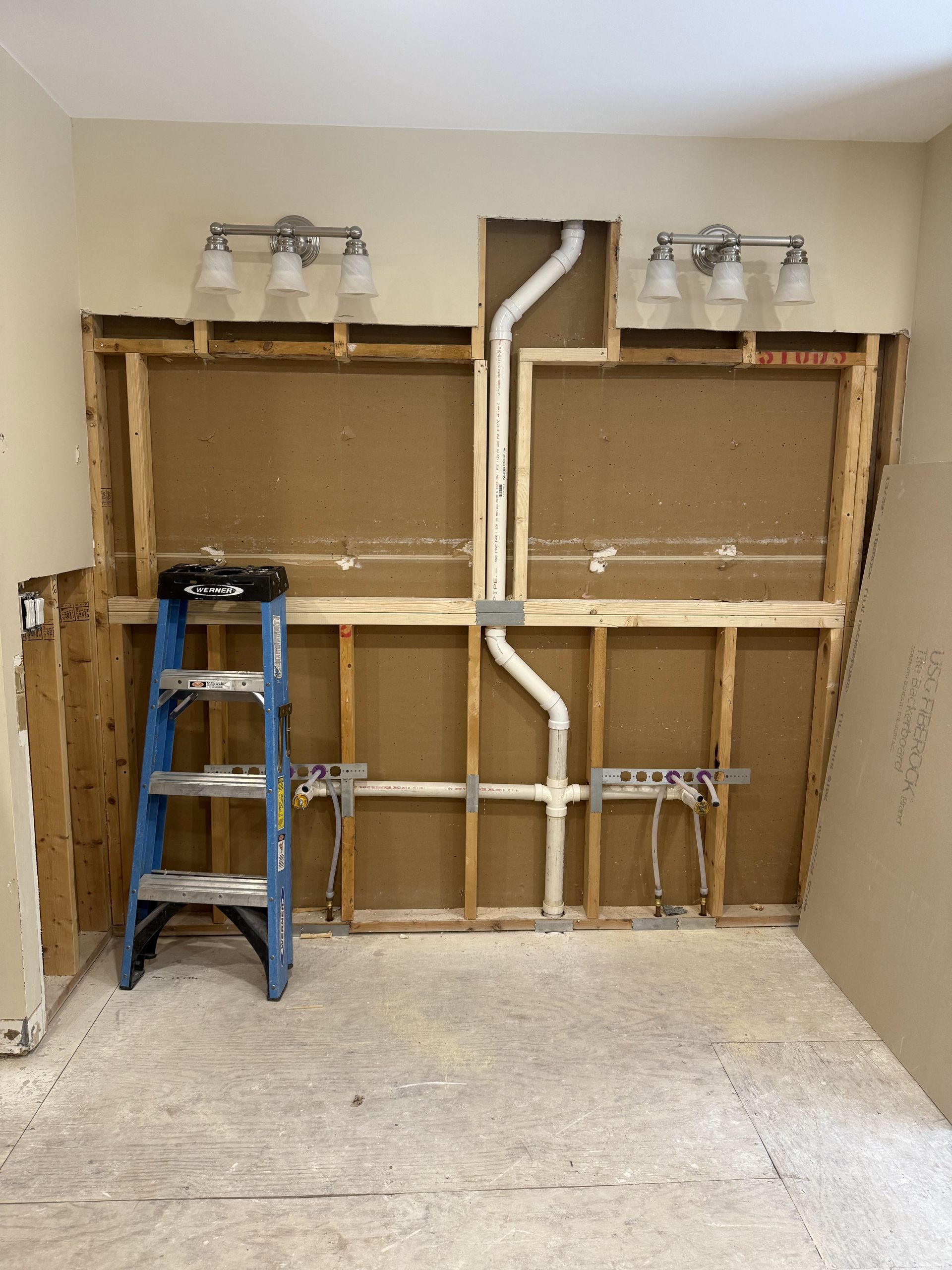Bathroom wall in renovation, exposed studs, plumbing visible, ladder, light fixtures.