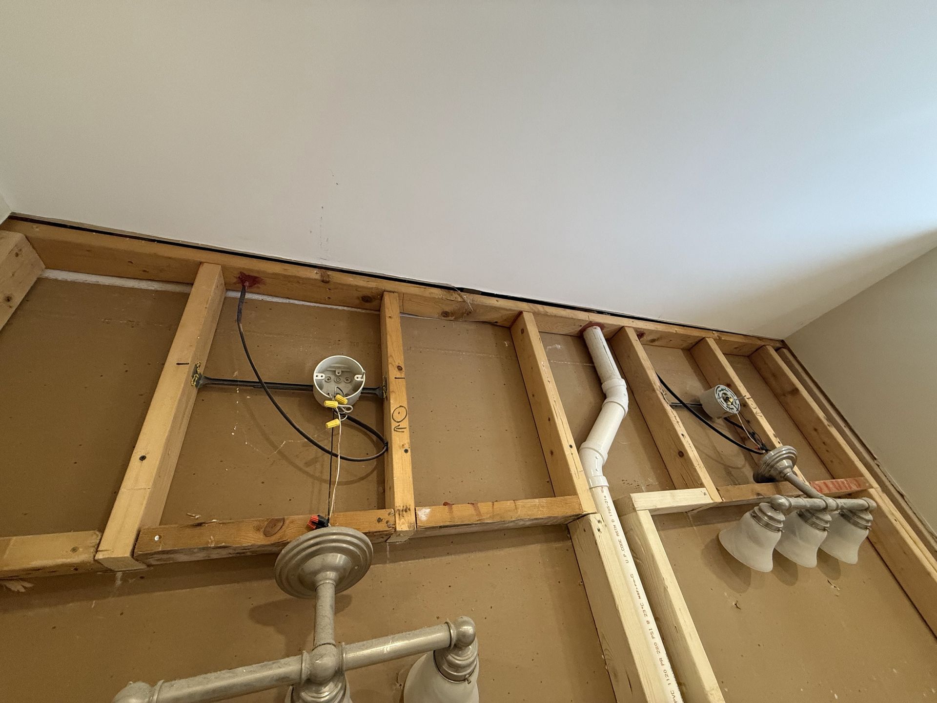 Exposed bathroom wall framing with electrical wiring, plumbing, and light fixtures.