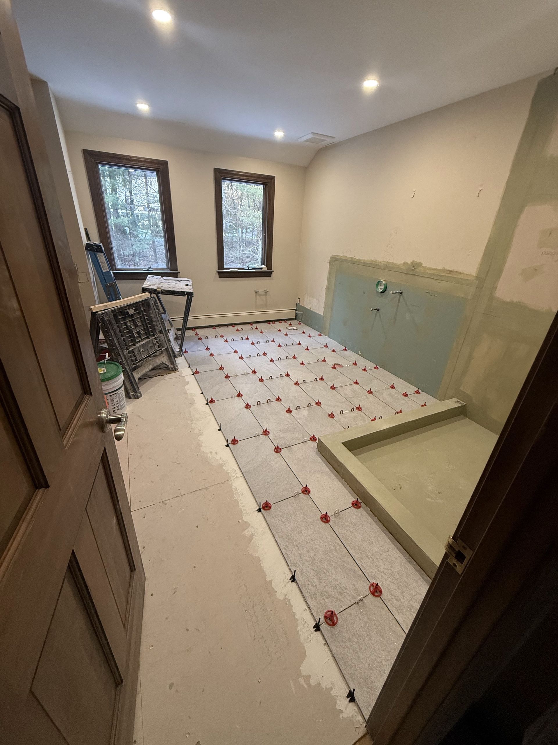 Bathroom renovation in progress; tiles laid out, unfinished walls, doorway visible, windows in background.