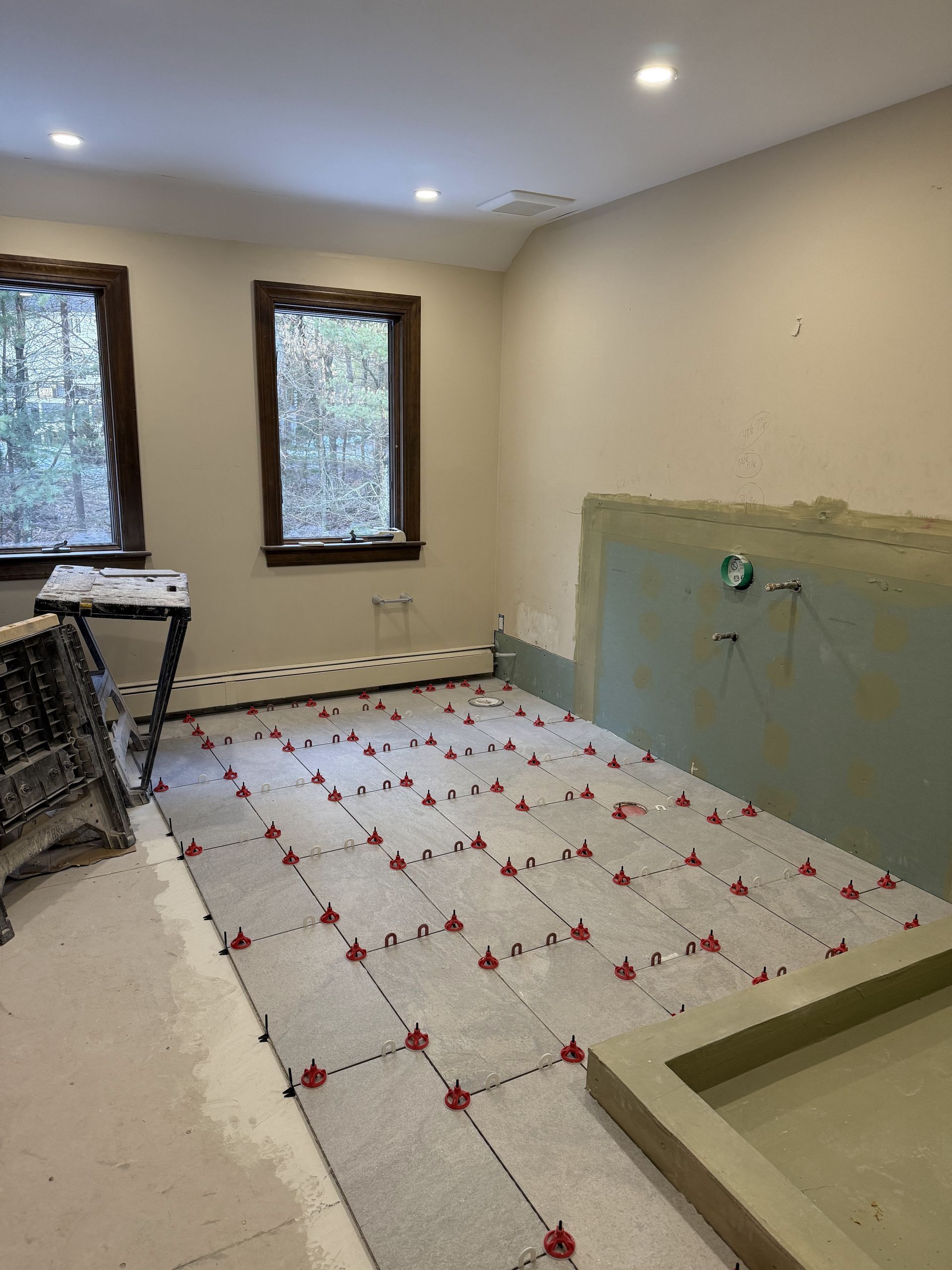 Bathroom under construction; tiled floor with leveling system. Light beige walls, two windows, and shower base visible.