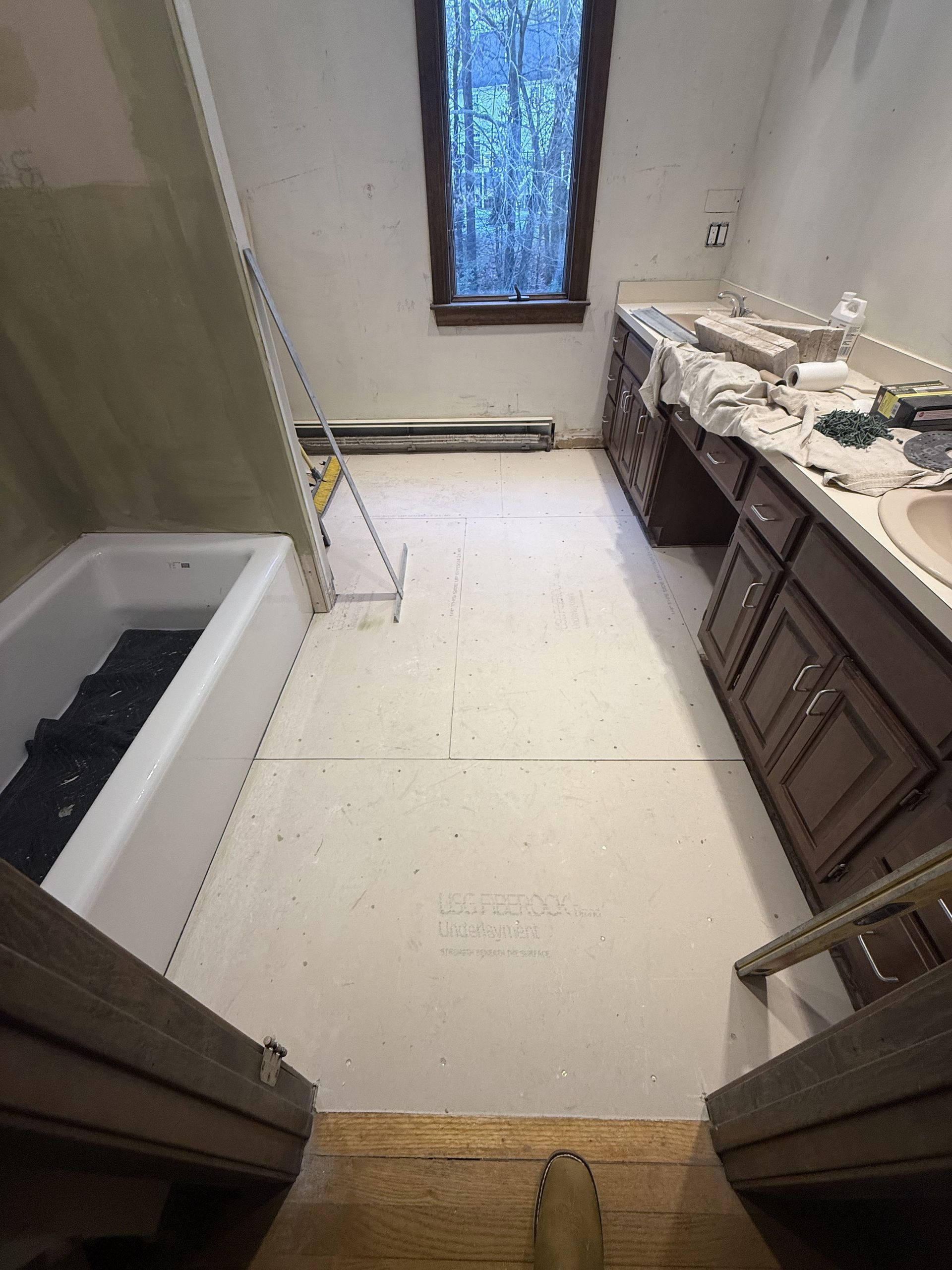 Bathroom undergoing renovation. White tub, vanity, and walls. Floor is light-colored and unfinished. Window at far end.