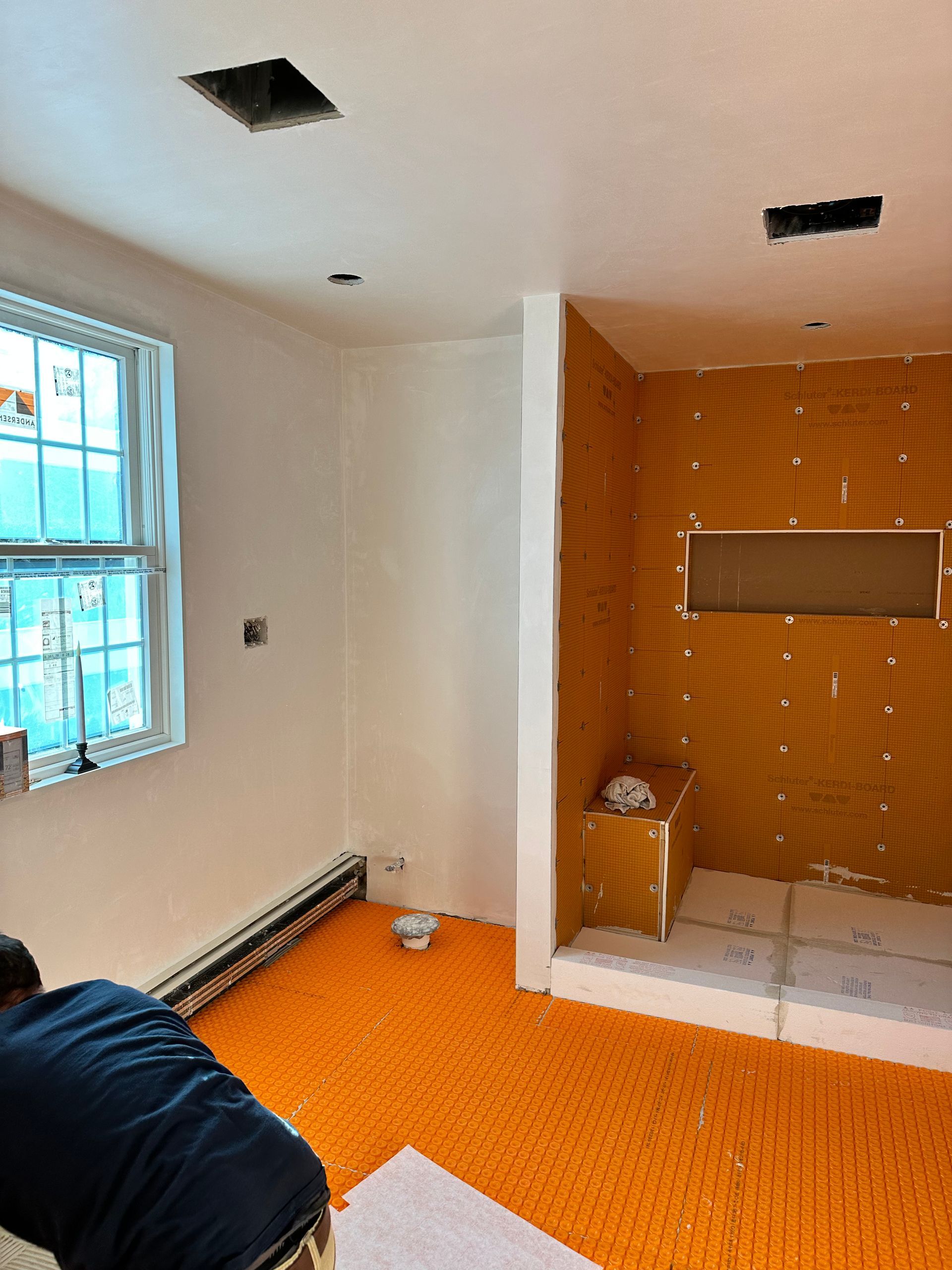 Bathroom renovation in progress; orange floor, shower stall, drywall, and a person working.