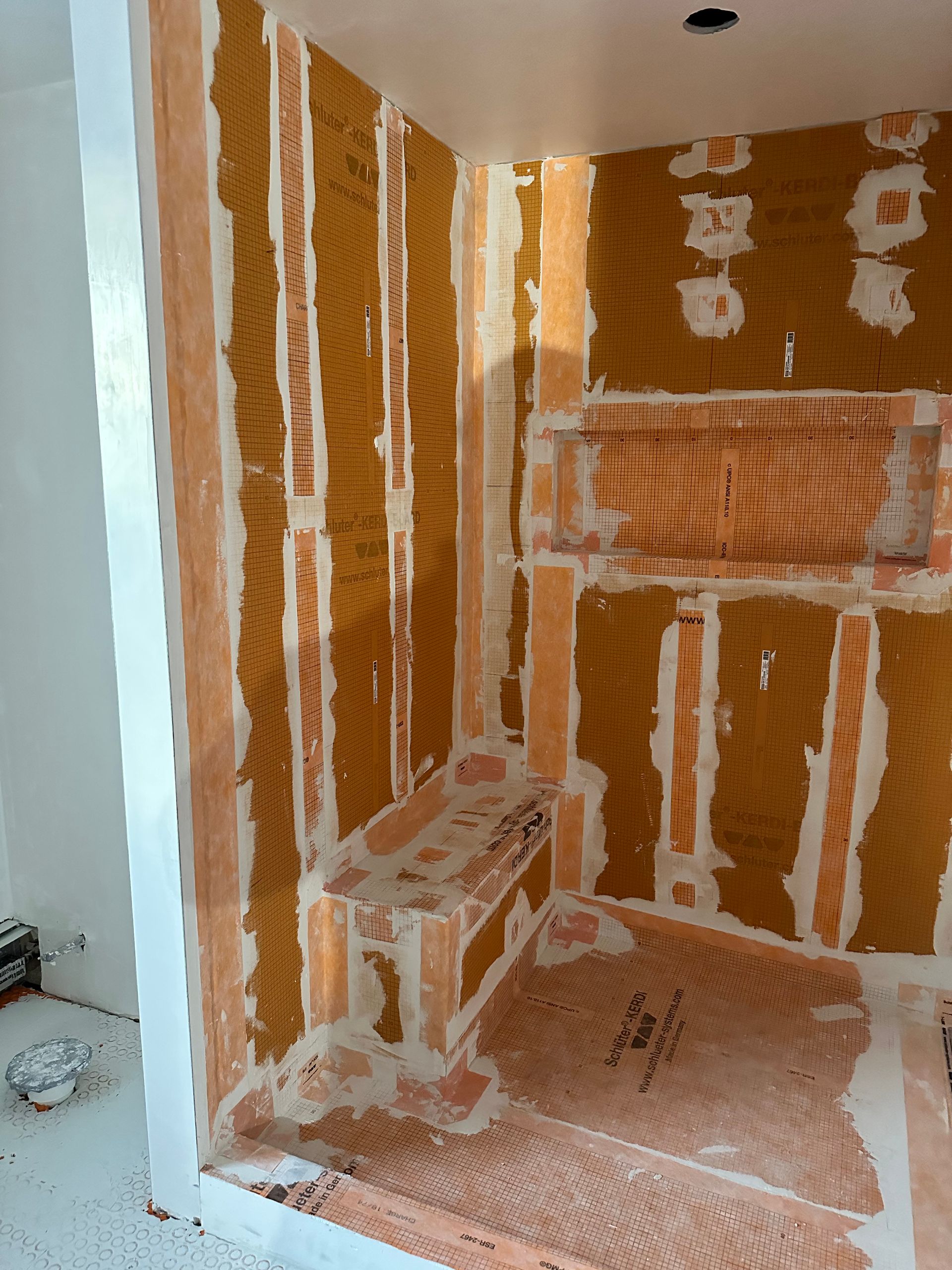 Shower stall under construction, orange backing with white mortar, includes a bench and shelf.