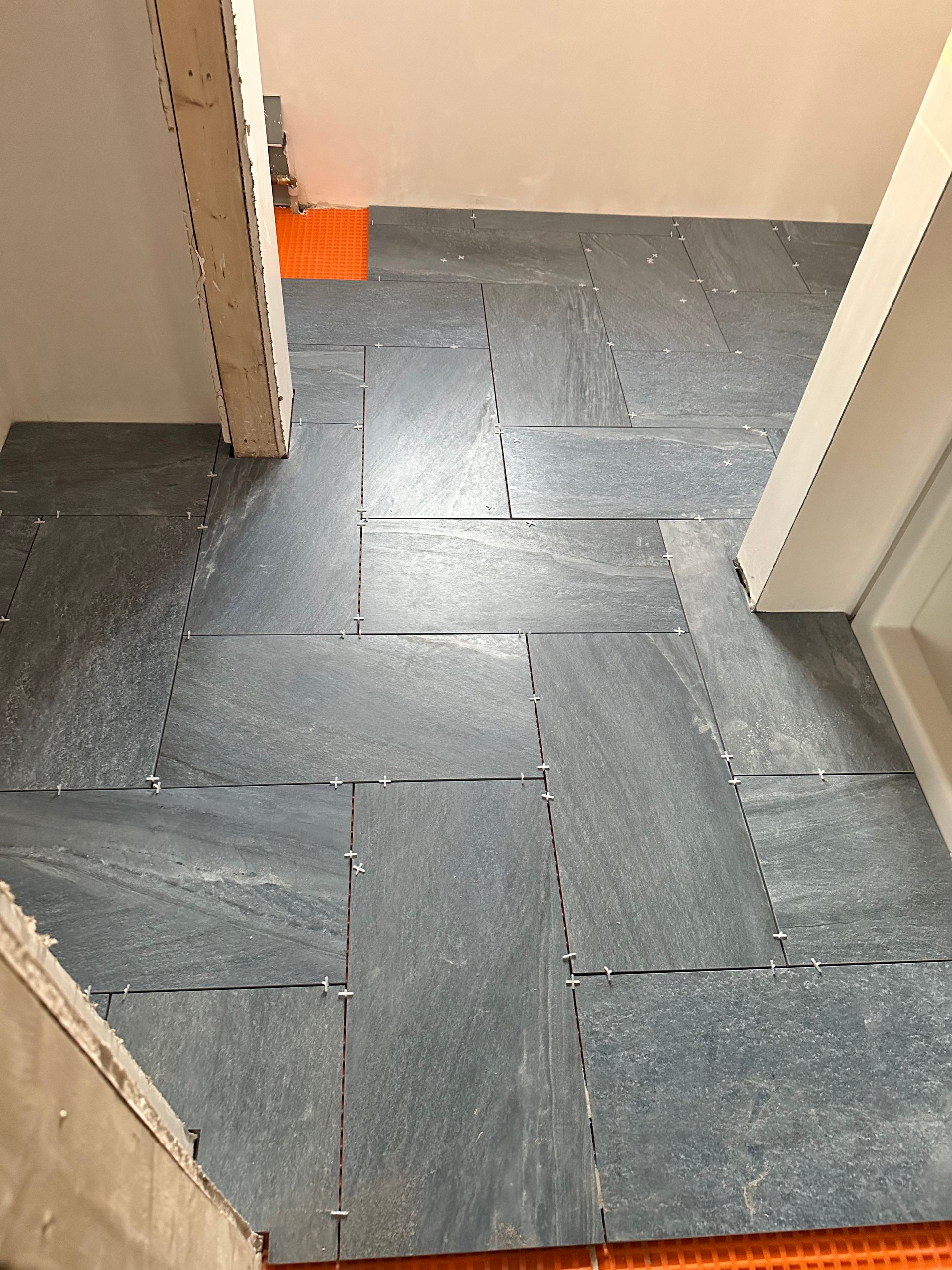 Gray tile floor installation in progress, orange leveling system in use, doorway visible.