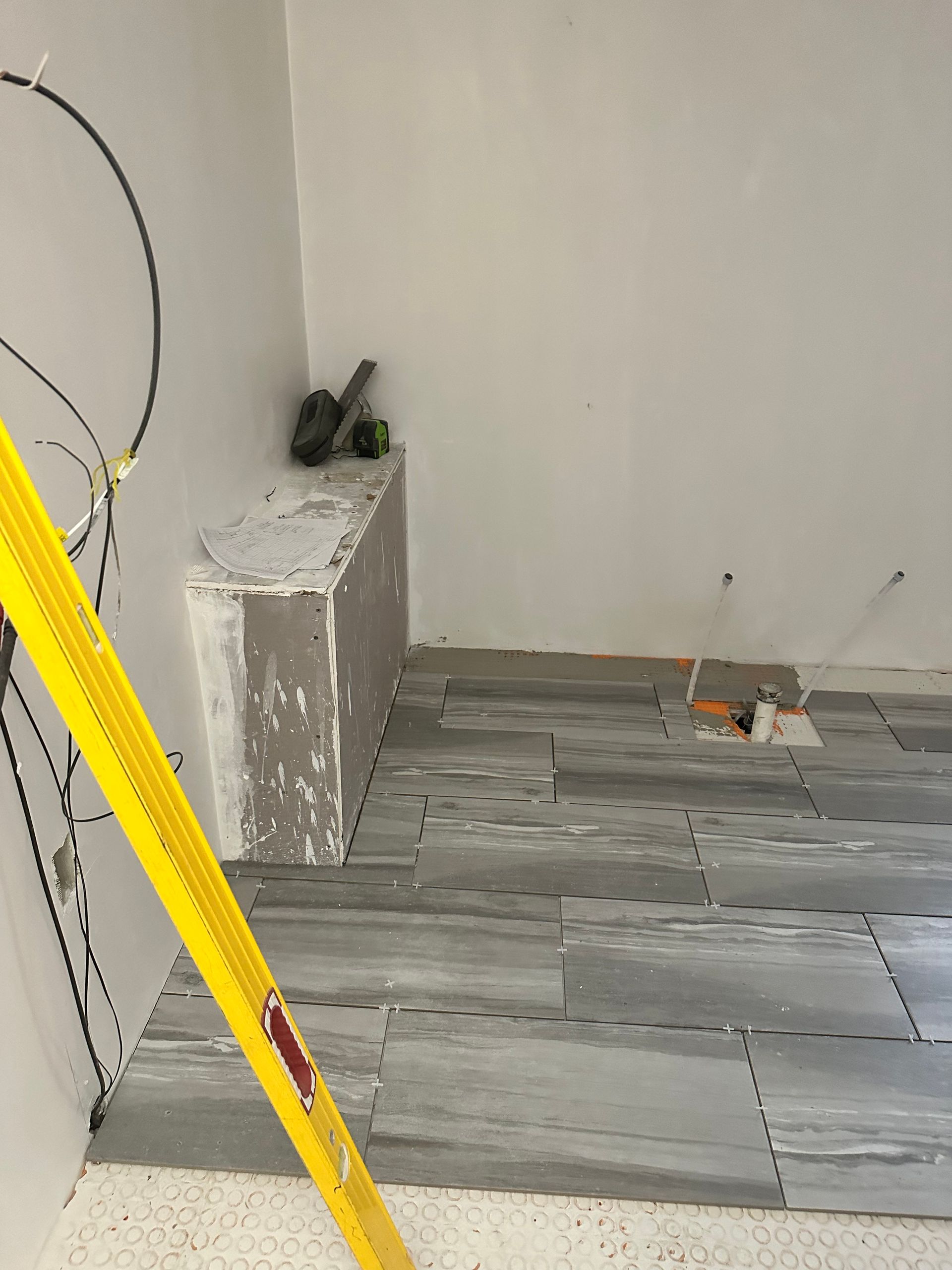 Bathroom under construction with gray stone-like tiles being laid on the floor. A yellow level rests on the tiles.