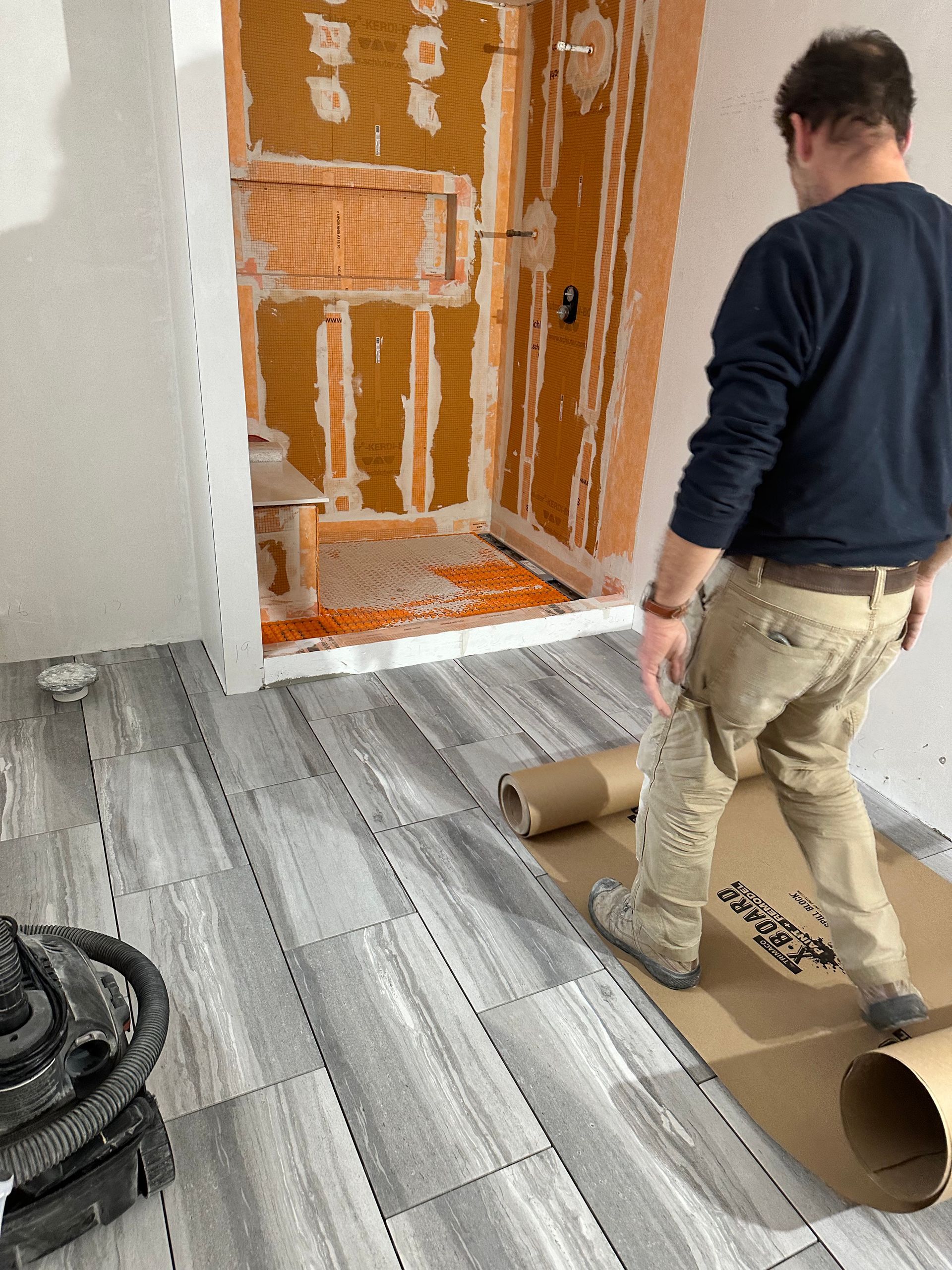 Man laying gray floor tiles in a bathroom under construction. Orange shower wall in the background.