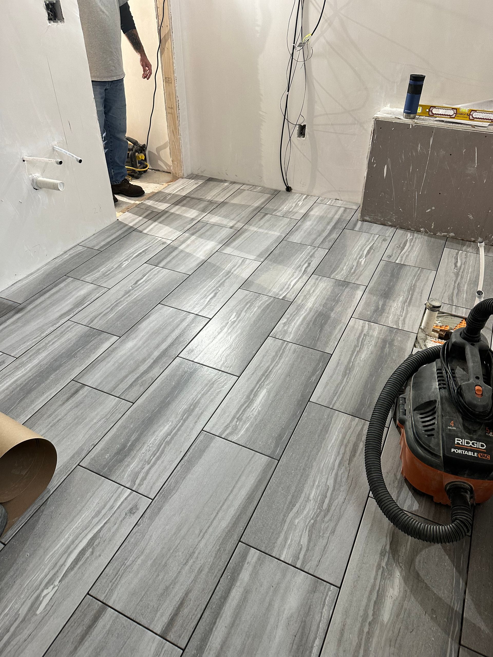 A room with gray wood-look tile flooring being installed; person standing, tools present.