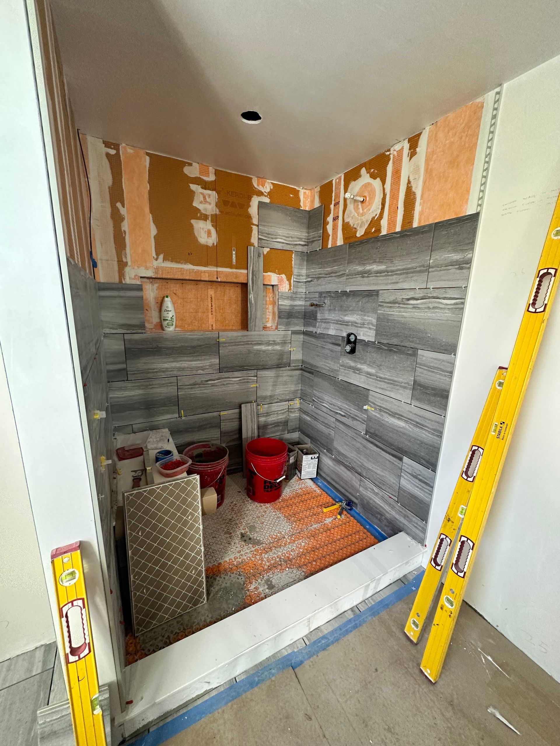 Shower under construction, gray tile installed on walls. Tools, materials, and a red bucket are in the unfinished space.