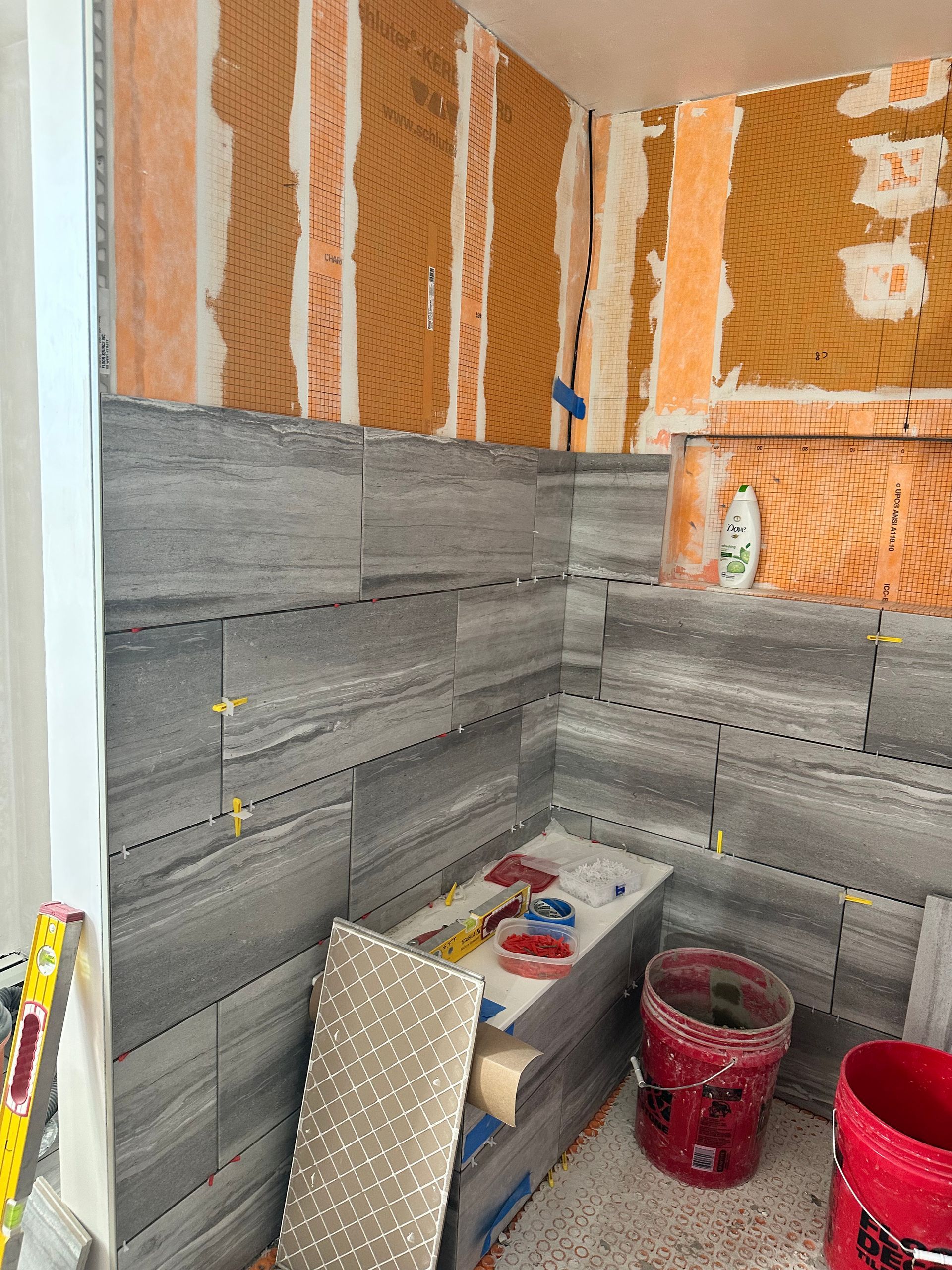 Bathroom under construction; gray tiles partially installed on the walls. Orange mesh and unfinished wall sections visible.