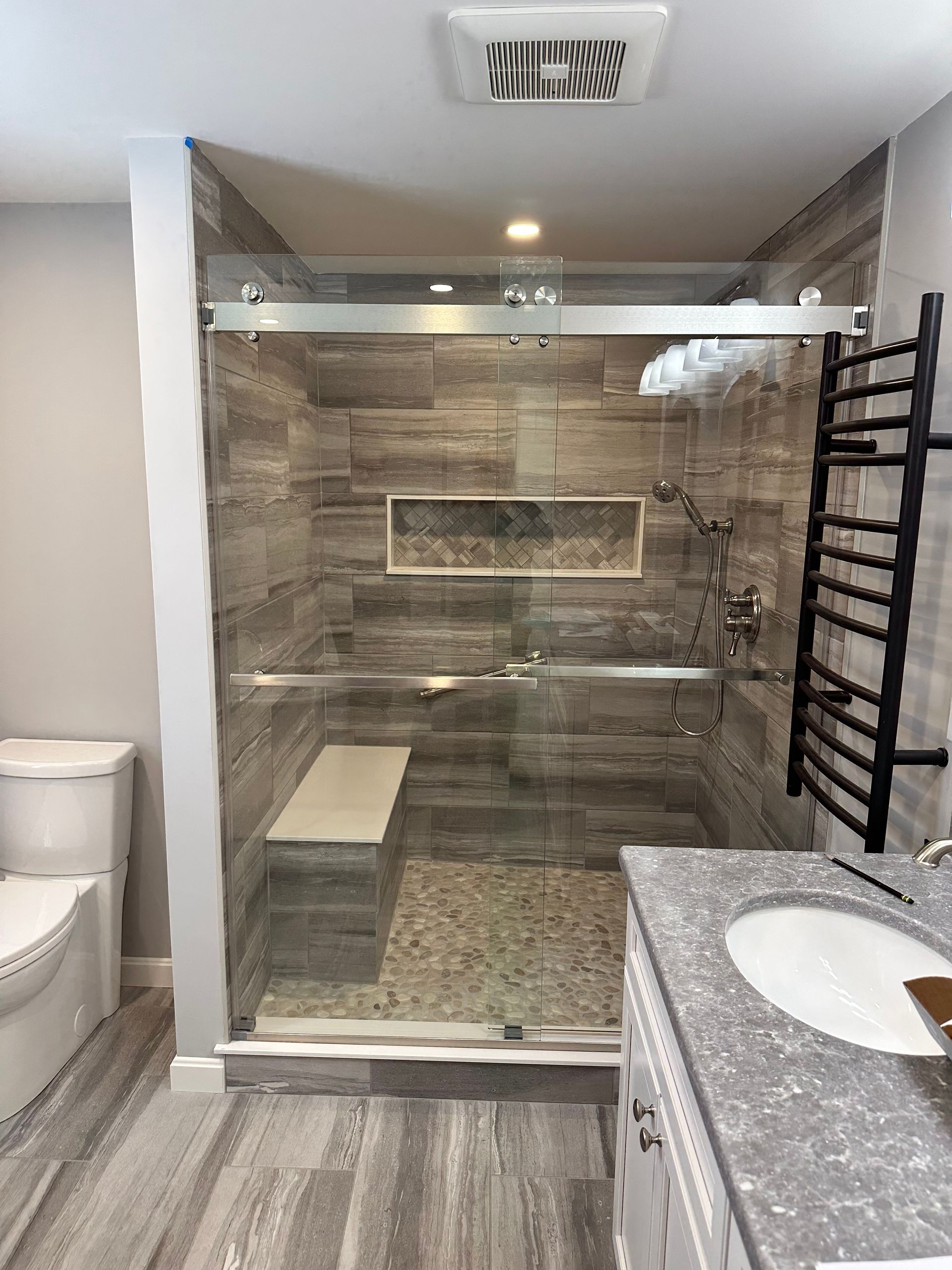 Modern bathroom with gray tiled shower, glass door, bench, and towel warmer.