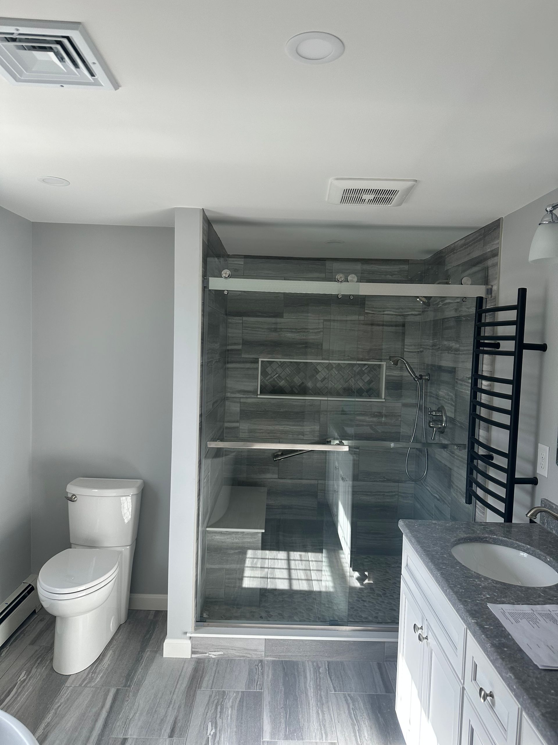 Gray bathroom with a glass shower, toilet, vanity, and heated towel rack.