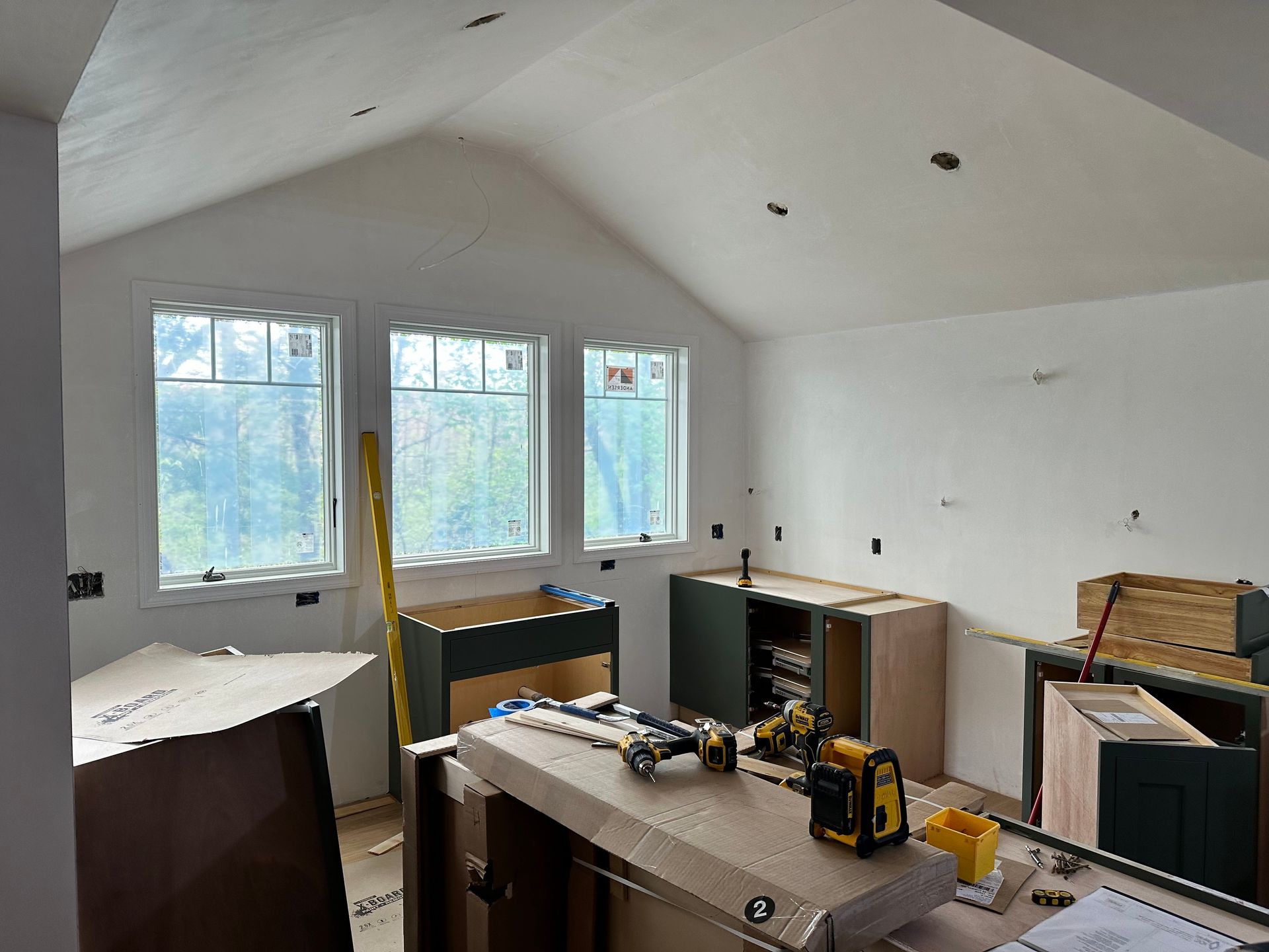 Kitchen under construction with cabinets, windows, and tools. White walls and vaulted ceiling.