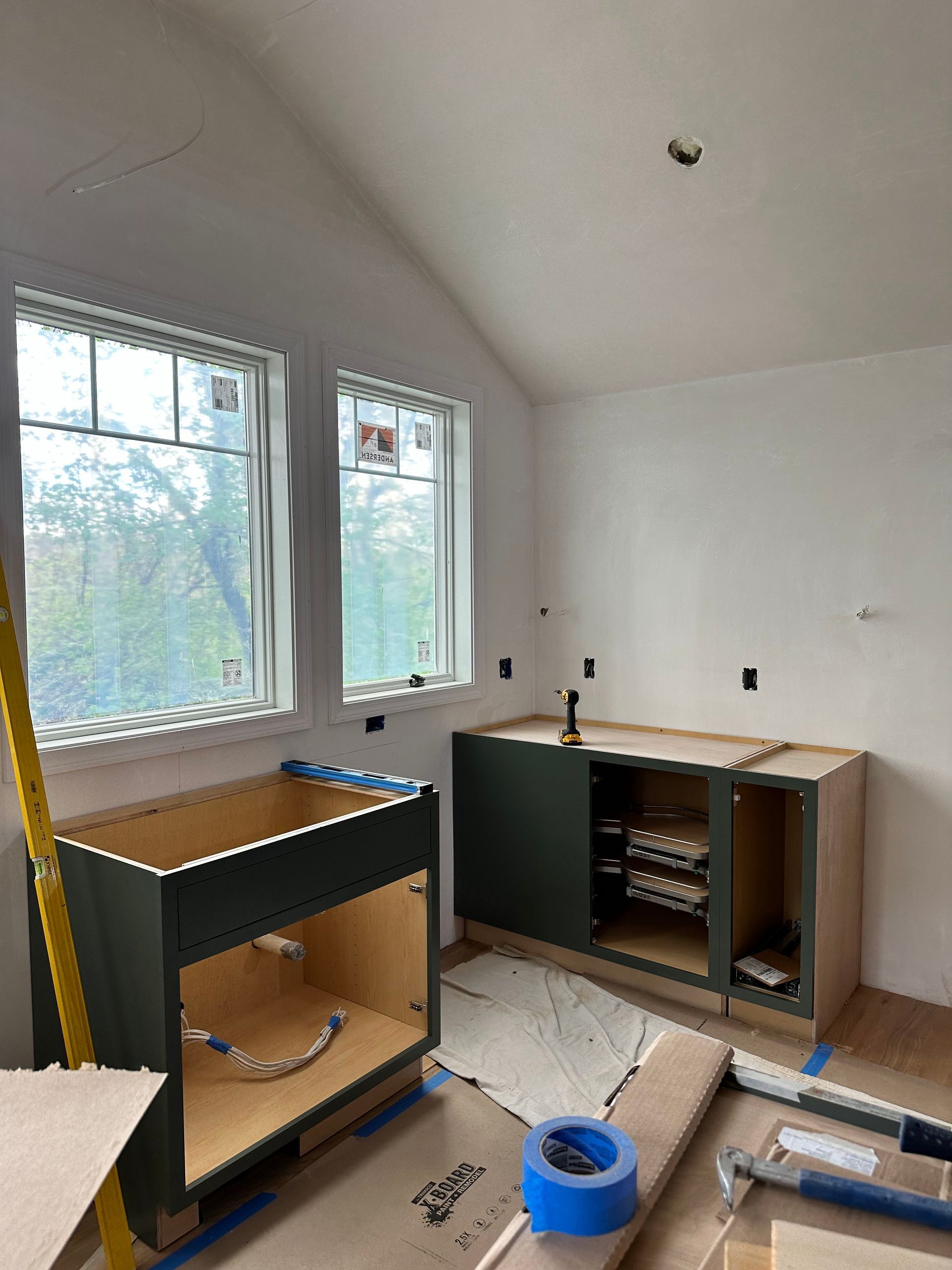 Kitchen cabinets in construction; green painted base cabinets; two windows on white walls.