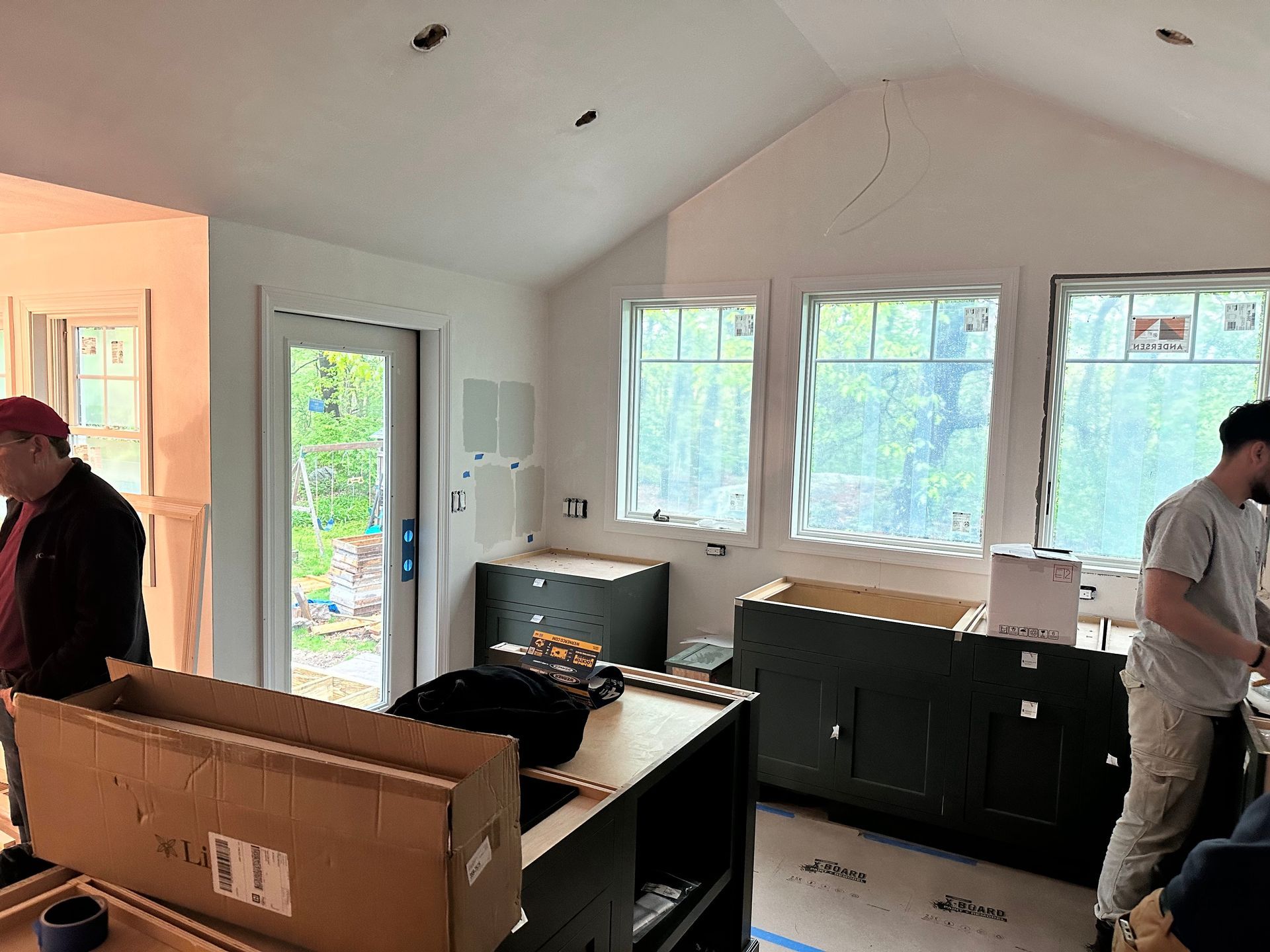 Interior of a kitchen under construction with cabinets, windows, and two men working.