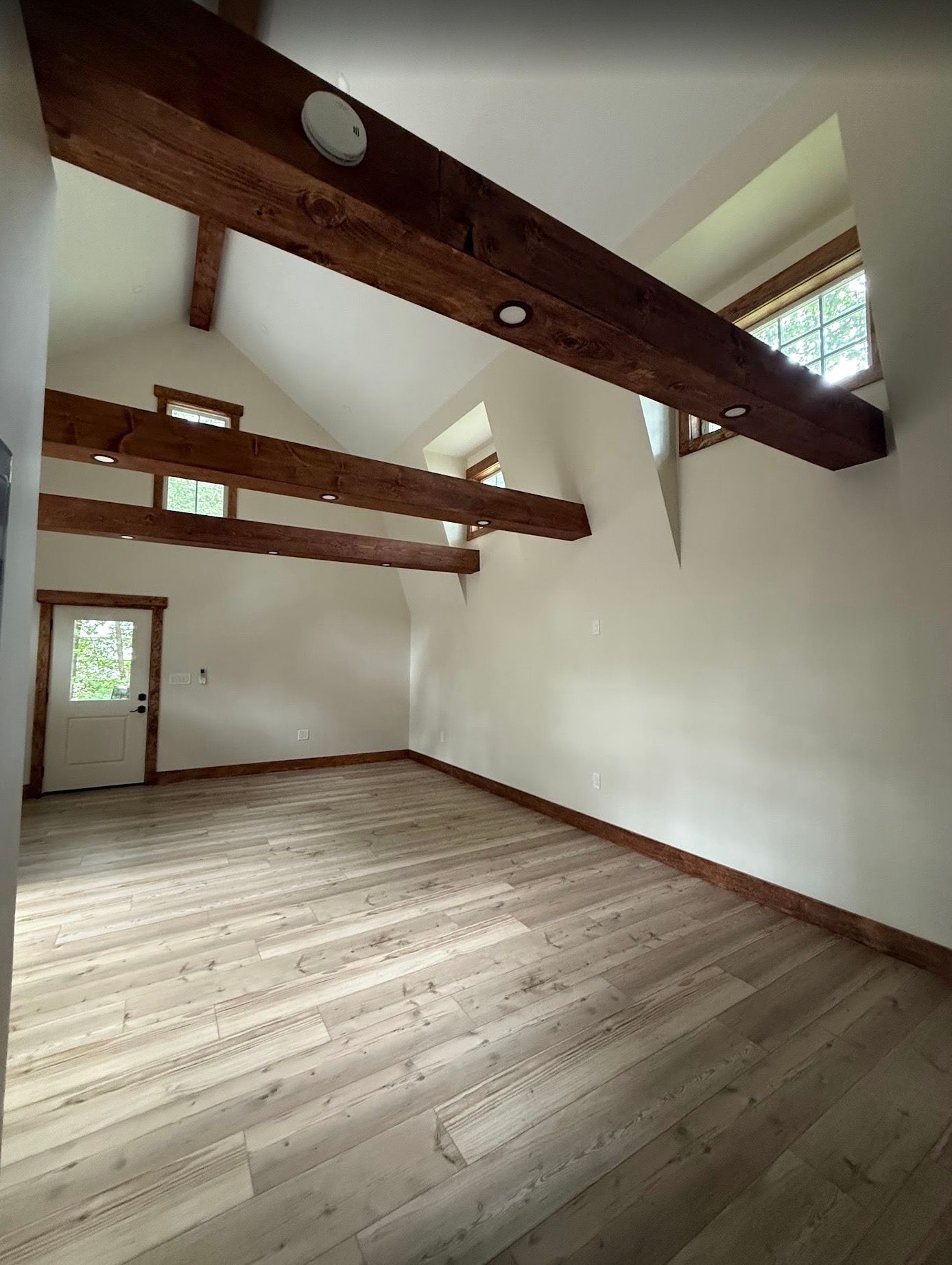 Empty room with light wood floors, white walls, and exposed wooden beams. Door on the left.