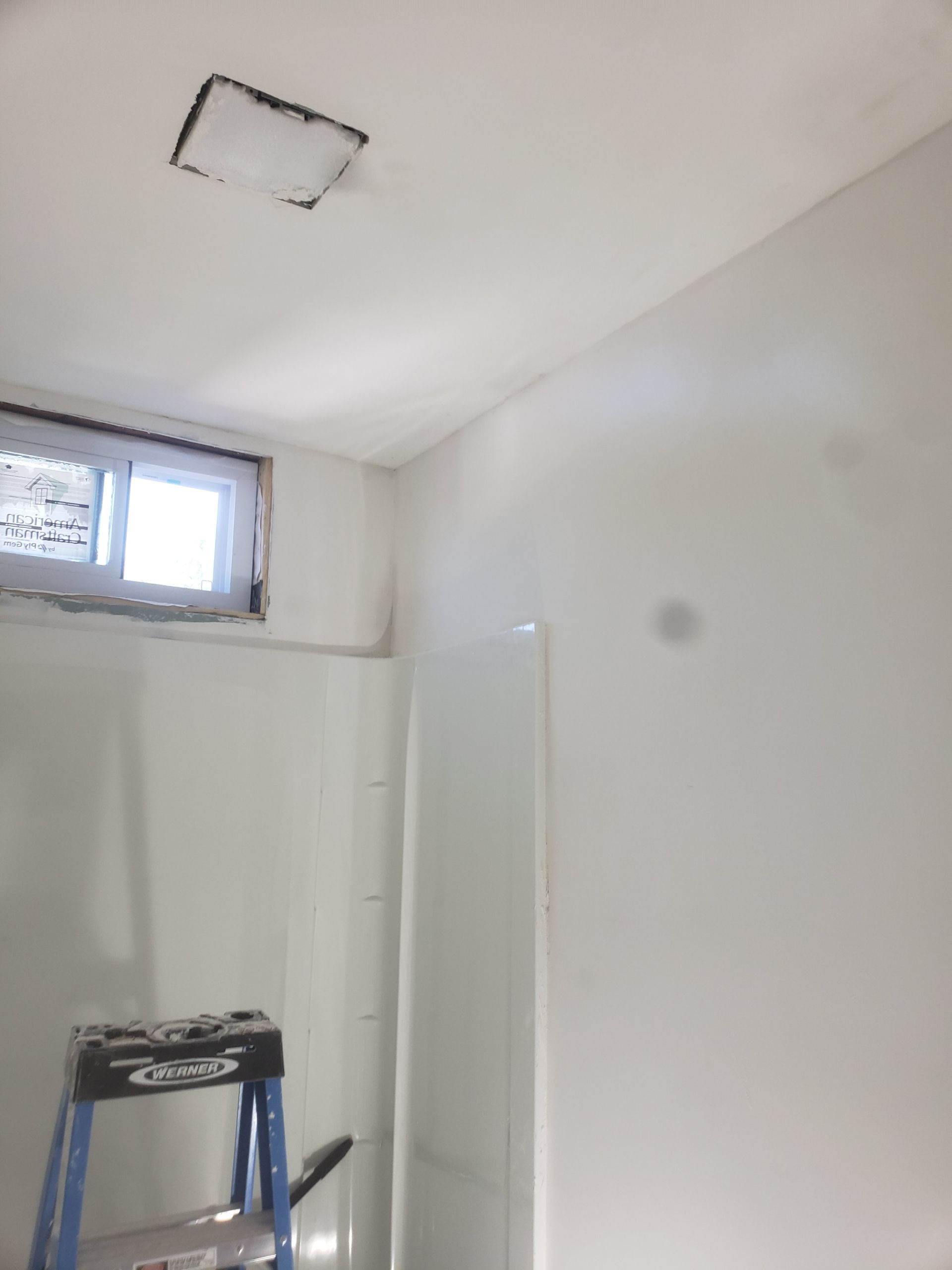 Bathroom with white walls and a shower unit; a window and a ceiling light are visible.