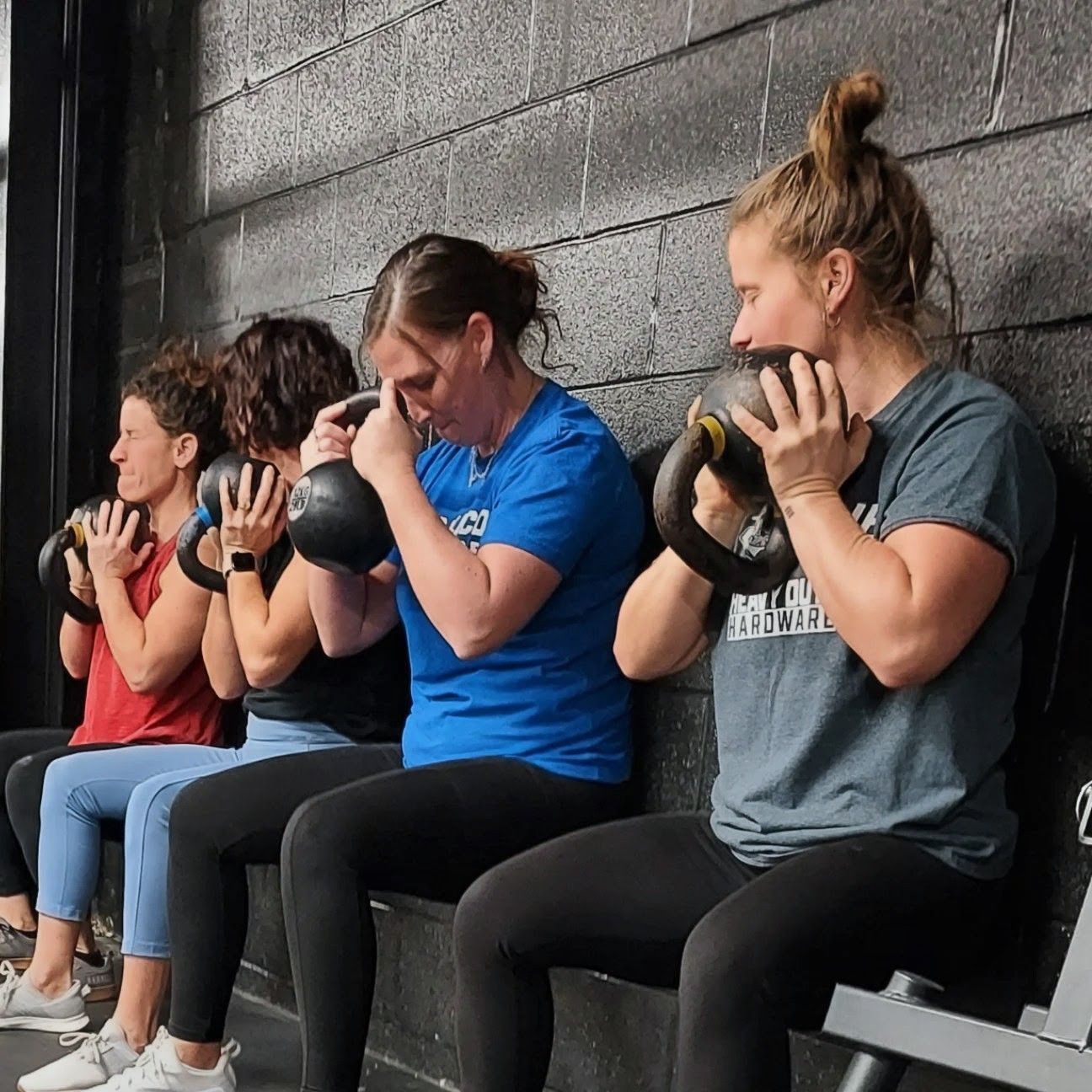 People in a fitness class hold kettlebells, seated against a black wall.