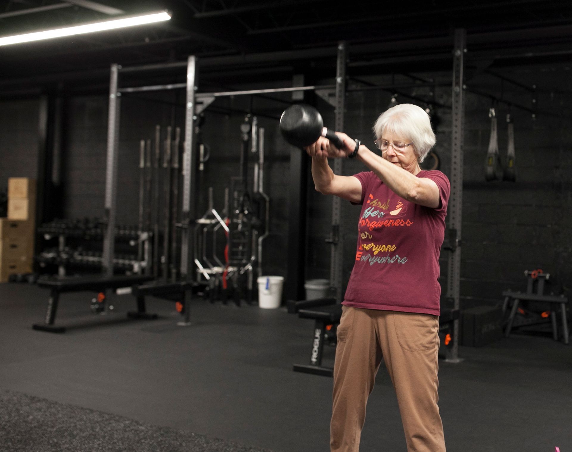 Woman in a gym swinging a black kettlebell. She is wearing a maroon shirt and brown pants.
