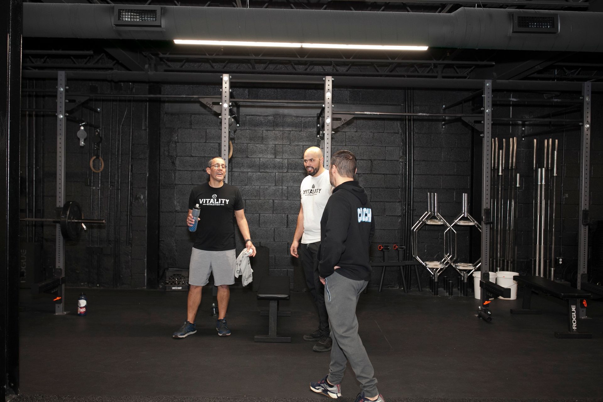 Three men in a gym, two talking, one holding a towel. Black bars, dark setting.