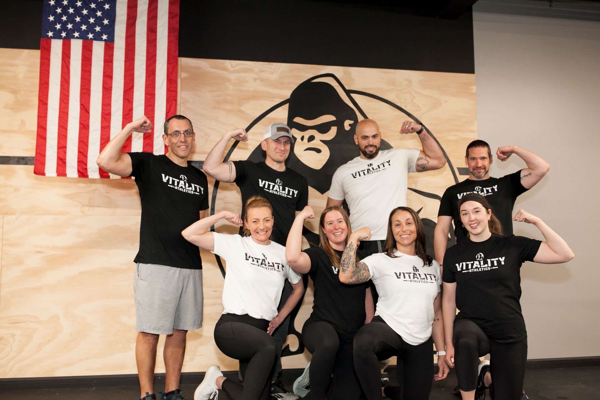 Group flexing in front of a gorilla logo and American flag. Gym setting, black and white shirts, smiling.