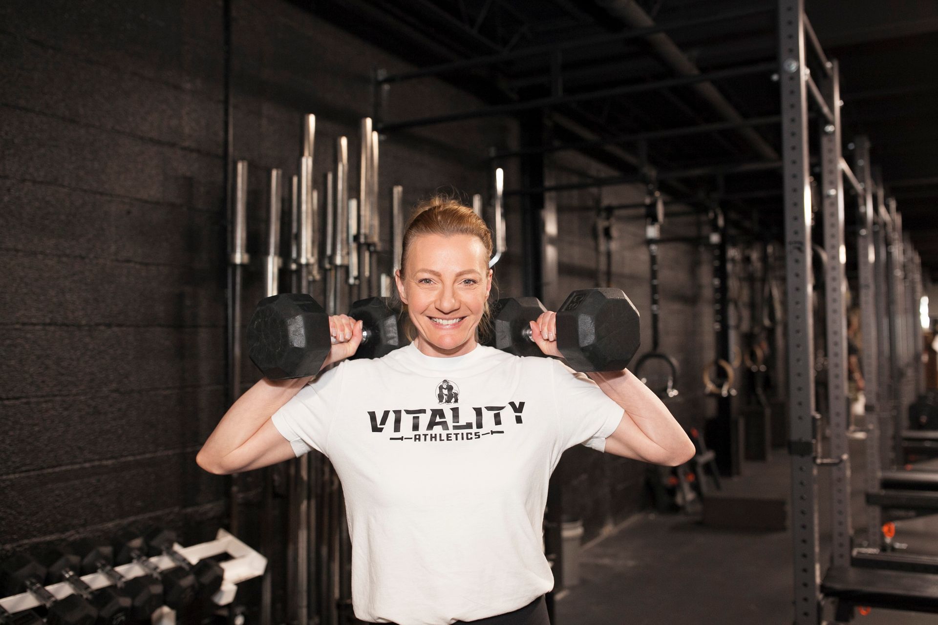 Woman in white t-shirt lifting dumbbells in a gym. Smiling, black wall background.