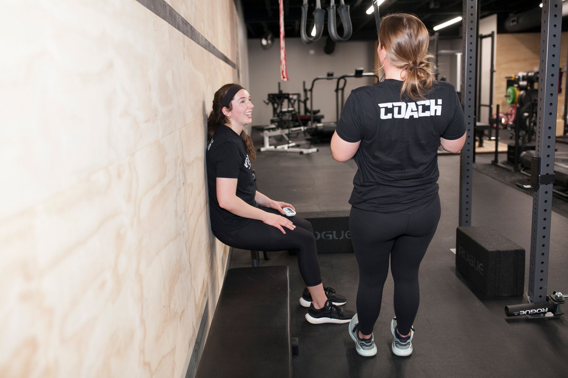 Woman doing wall sit exercise, supervised by a coach in a gym.