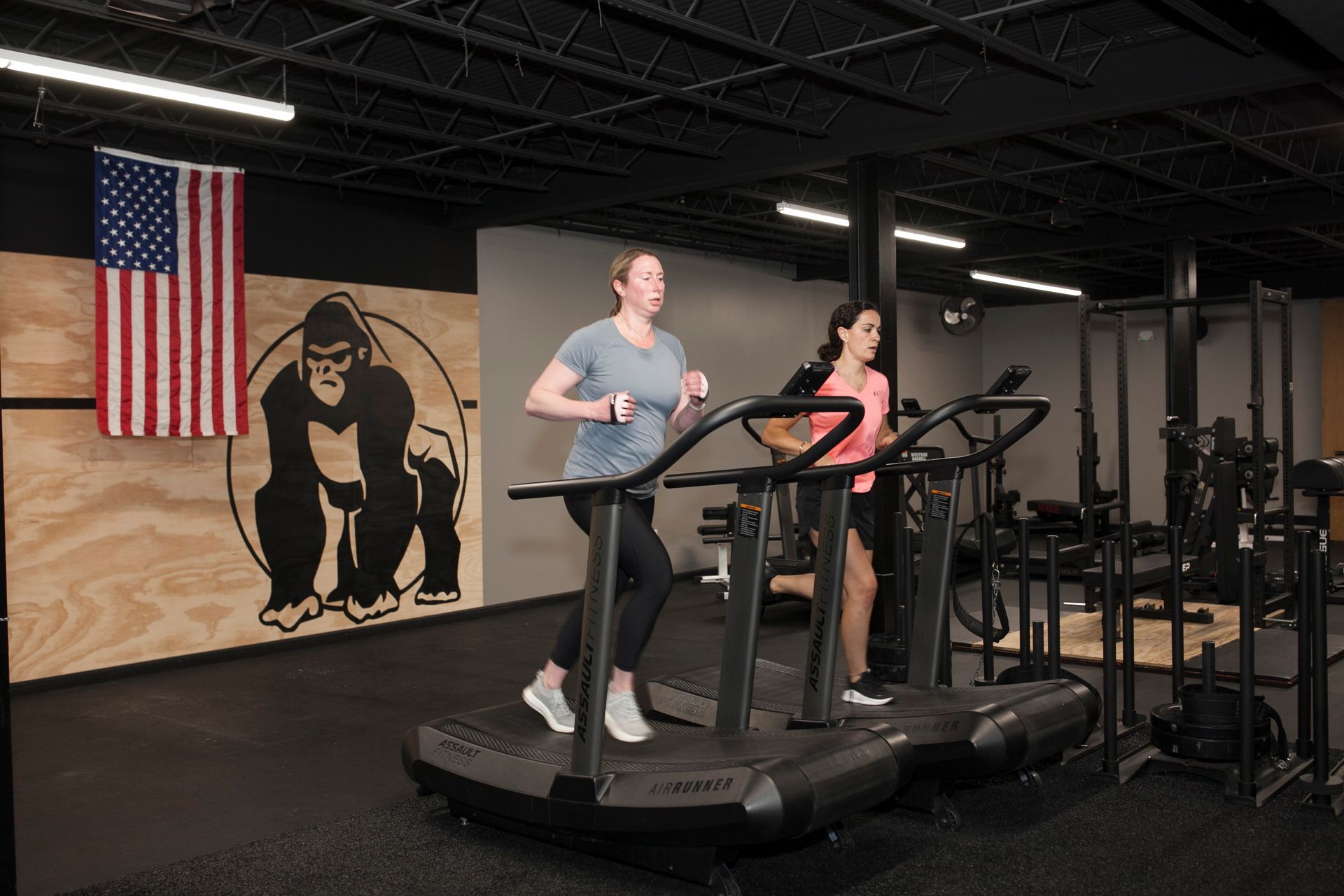 Two people running on curved treadmills in a gym with an American flag and gorilla logo.