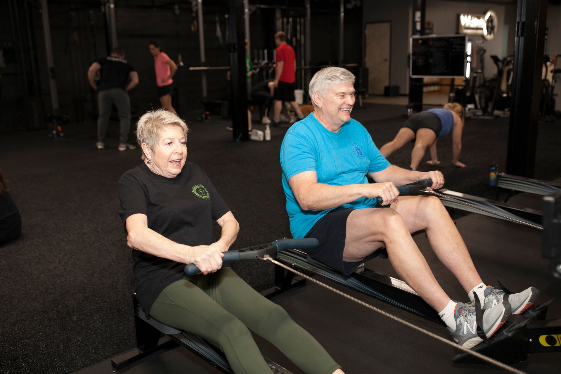 Two people rowing on machines at a gym. Others are exercising in the background.