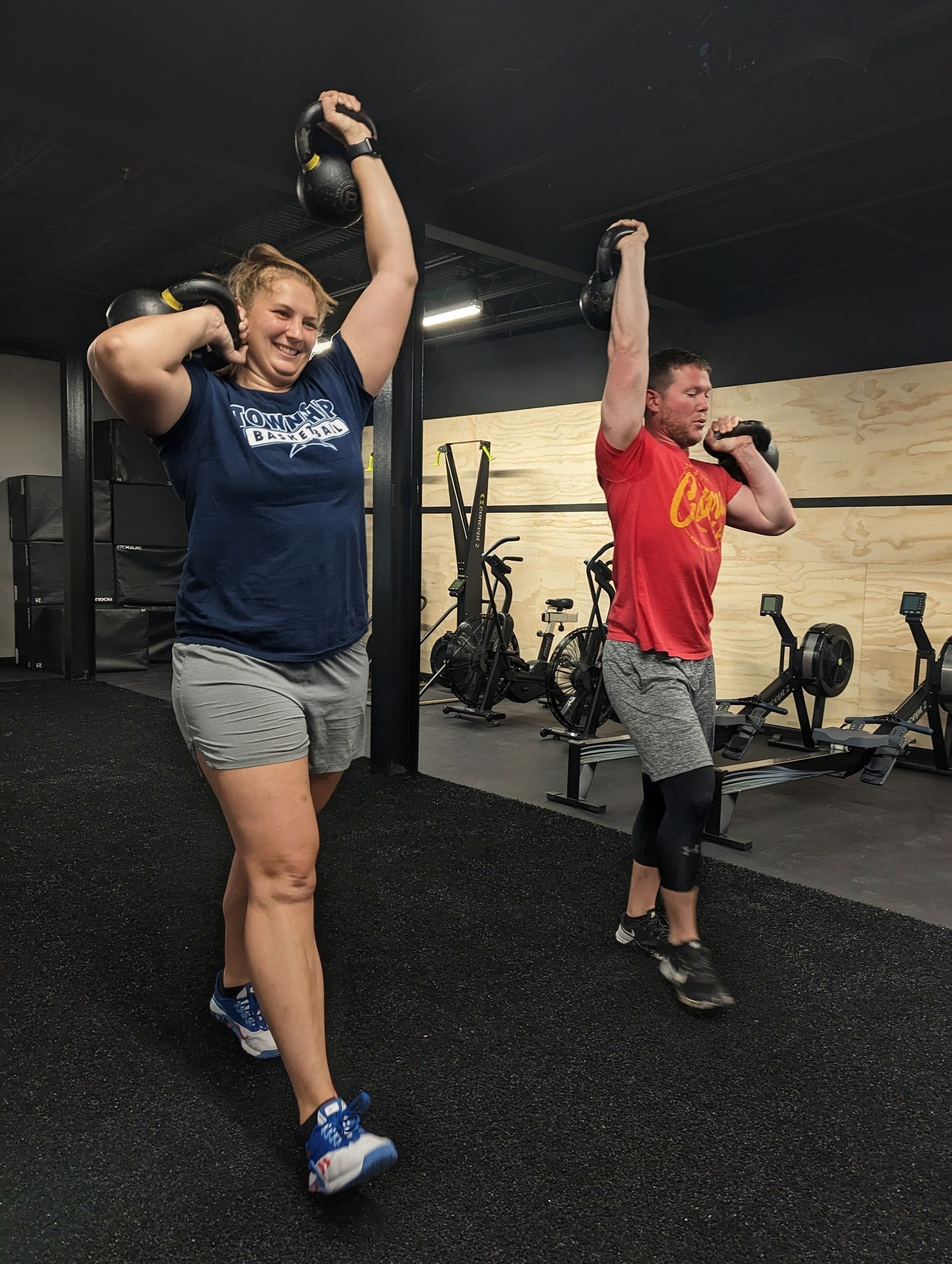Two people exercising in a gym, carrying kettlebells overhead. Black floor, neutral background.