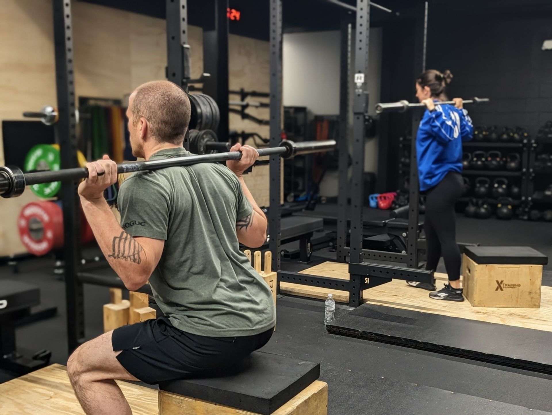 Man in gym squats with barbell on box. Woman squats behind, barbell on back.