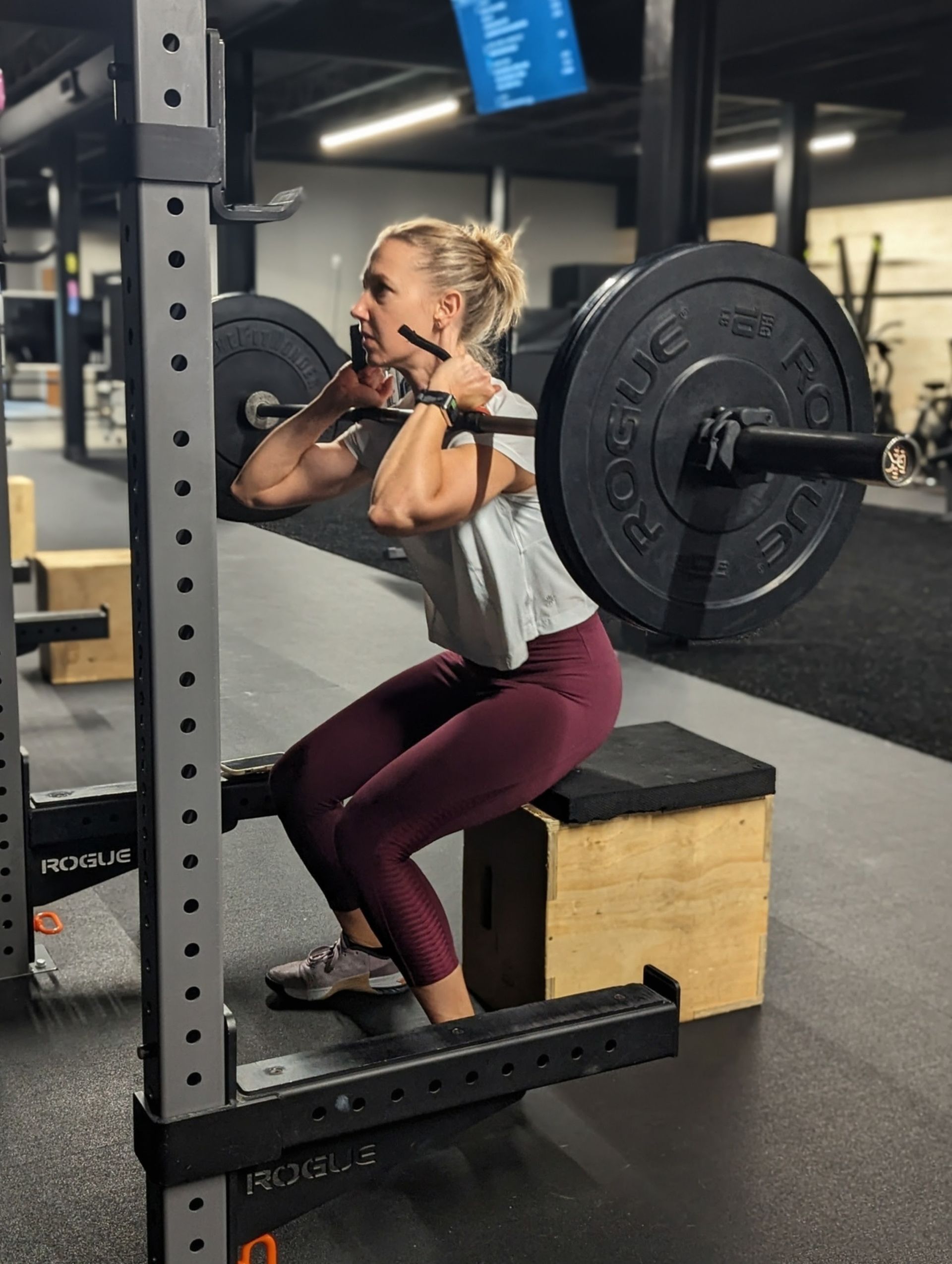 Woman performing a barbell front squat, using a box, at a gym. She wears maroon leggings and gray shirt.