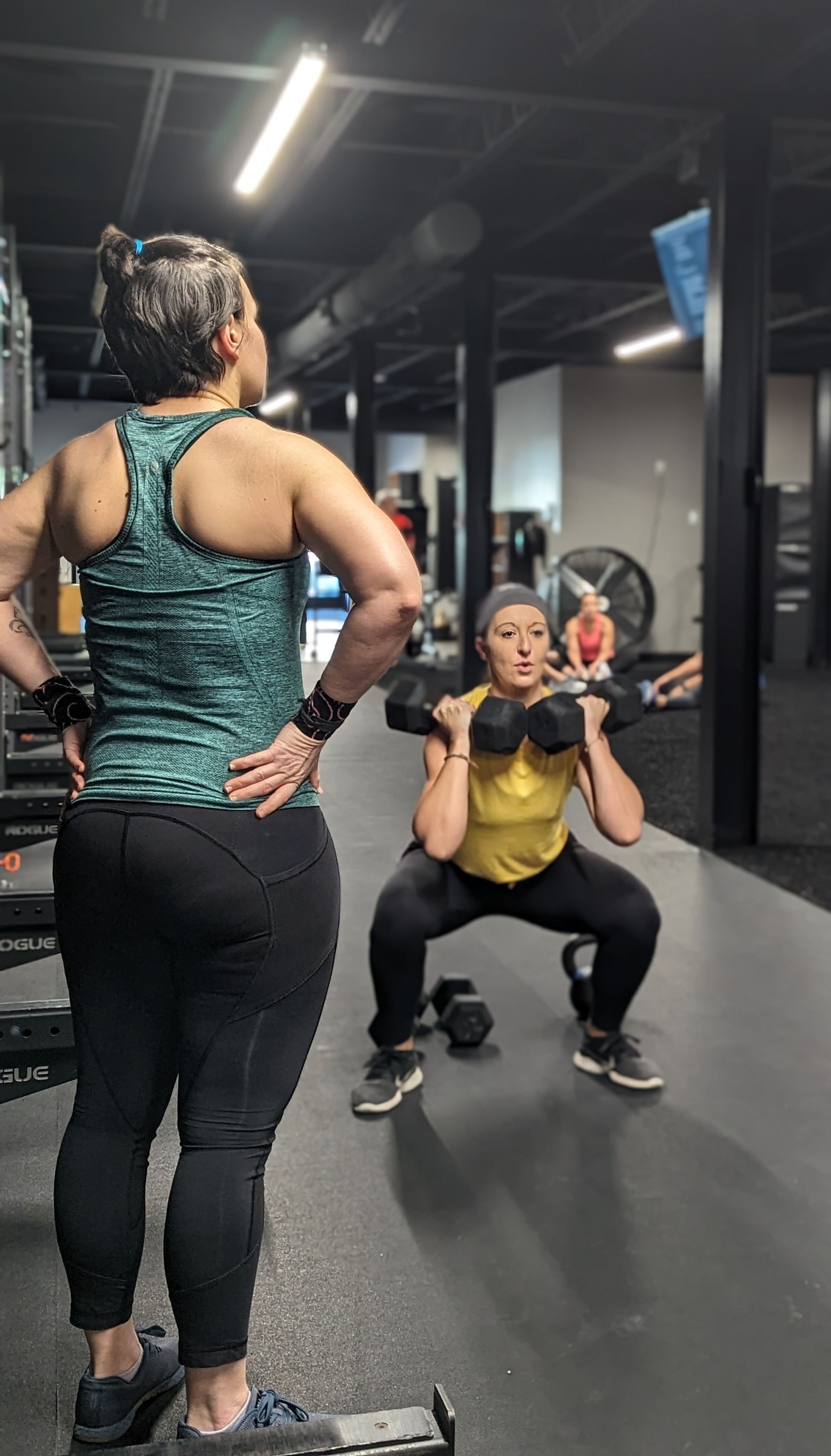 Woman squats with weight, watched by another in a gym.