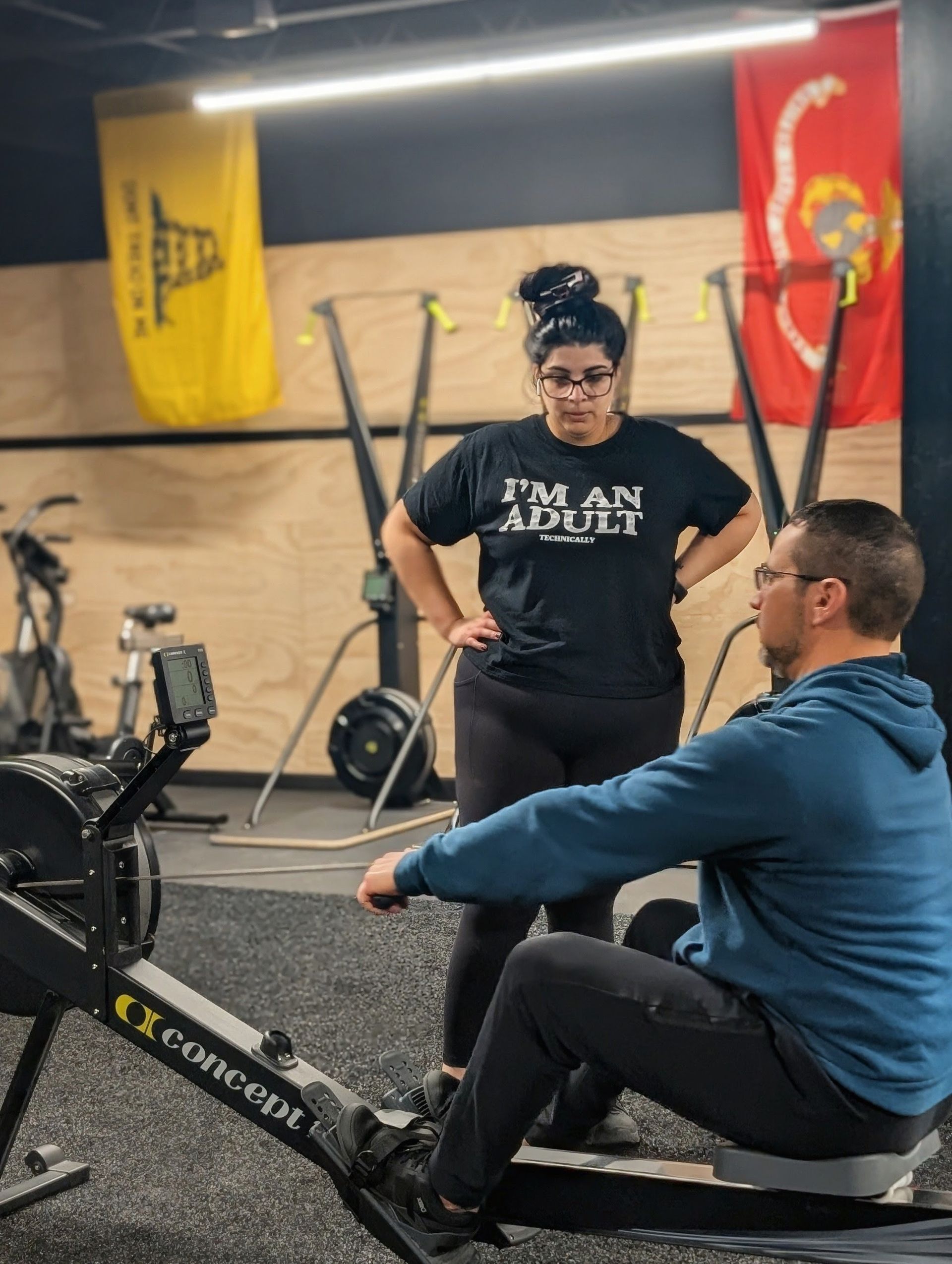 A woman coaches a man on a rowing machine in a gym. She wears a