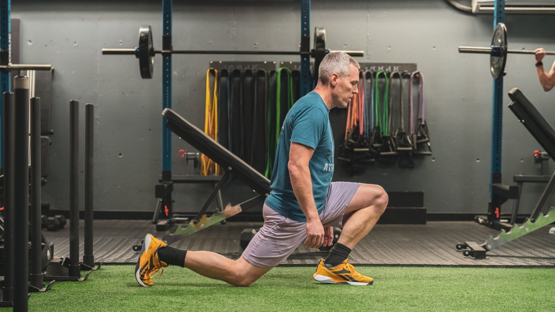 Man in a gym, performing a lunge. He has gray hair and is wearing workout attire; gray shorts, a blue shirt, and yellow sneakers.