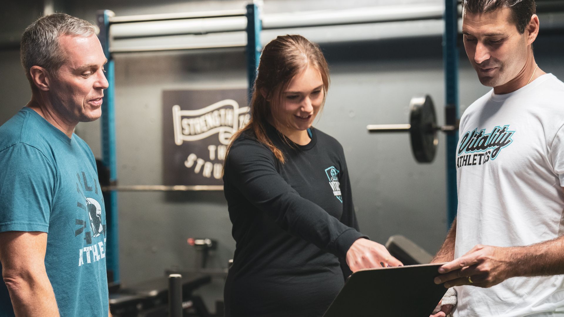 Three people in gym discuss document: woman points, men look. Weightlifting equipment in background.