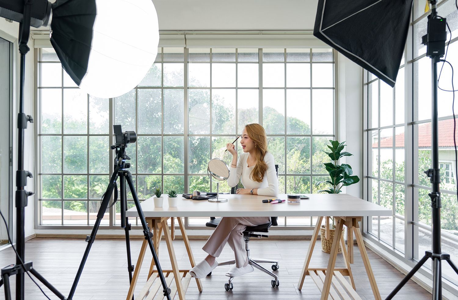 A woman is sitting at a desk in front of a camera applying makeup.