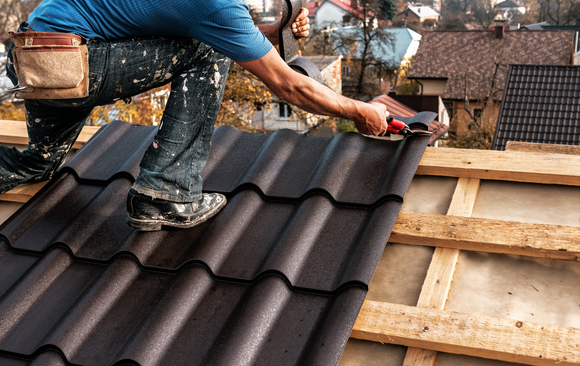 Roofer installing dark metal roof tiles on a wooden framework outdoors, with houses in background.