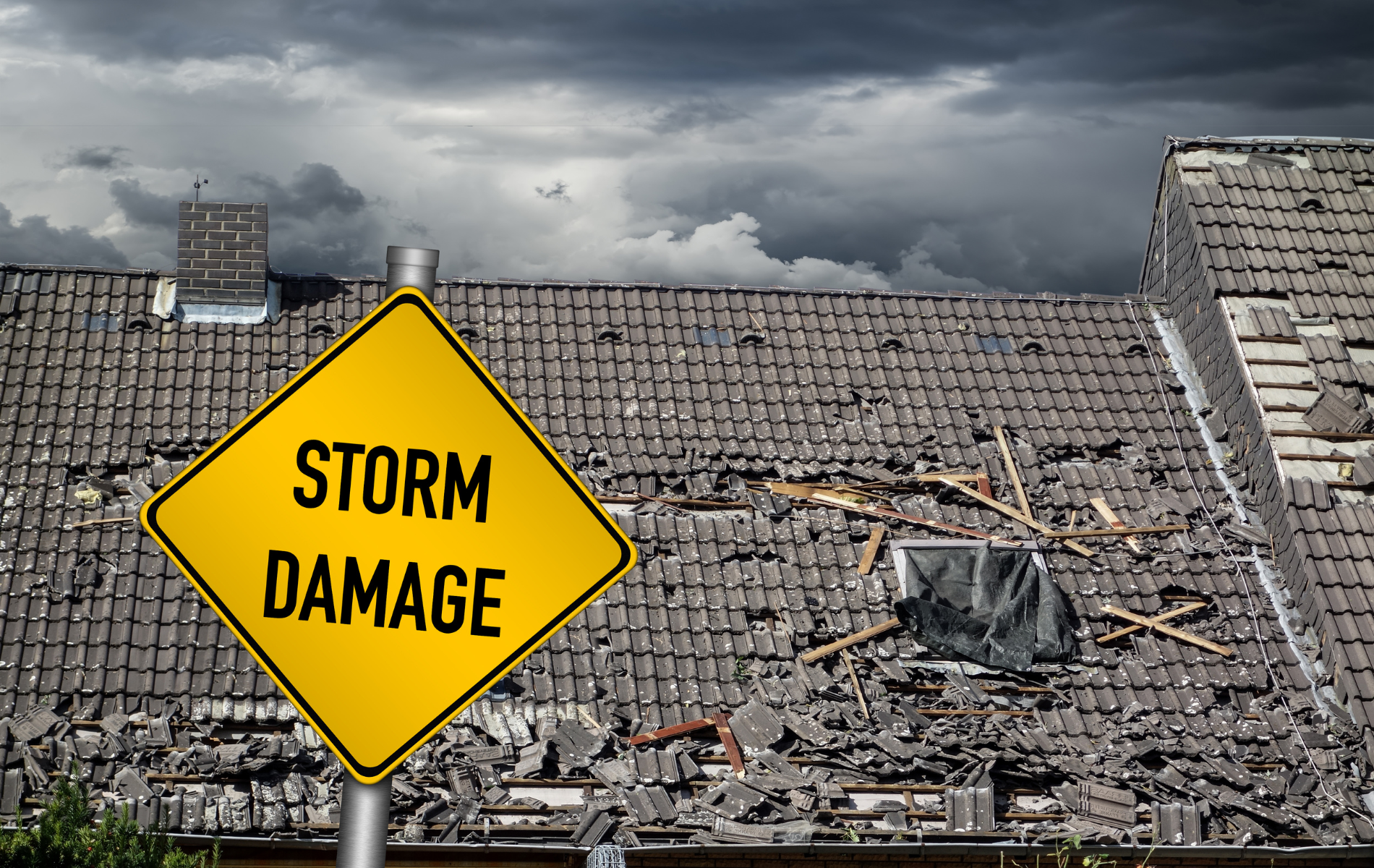 Yellow storm damage sign in front of a damaged house roof under a dark, stormy sky.
