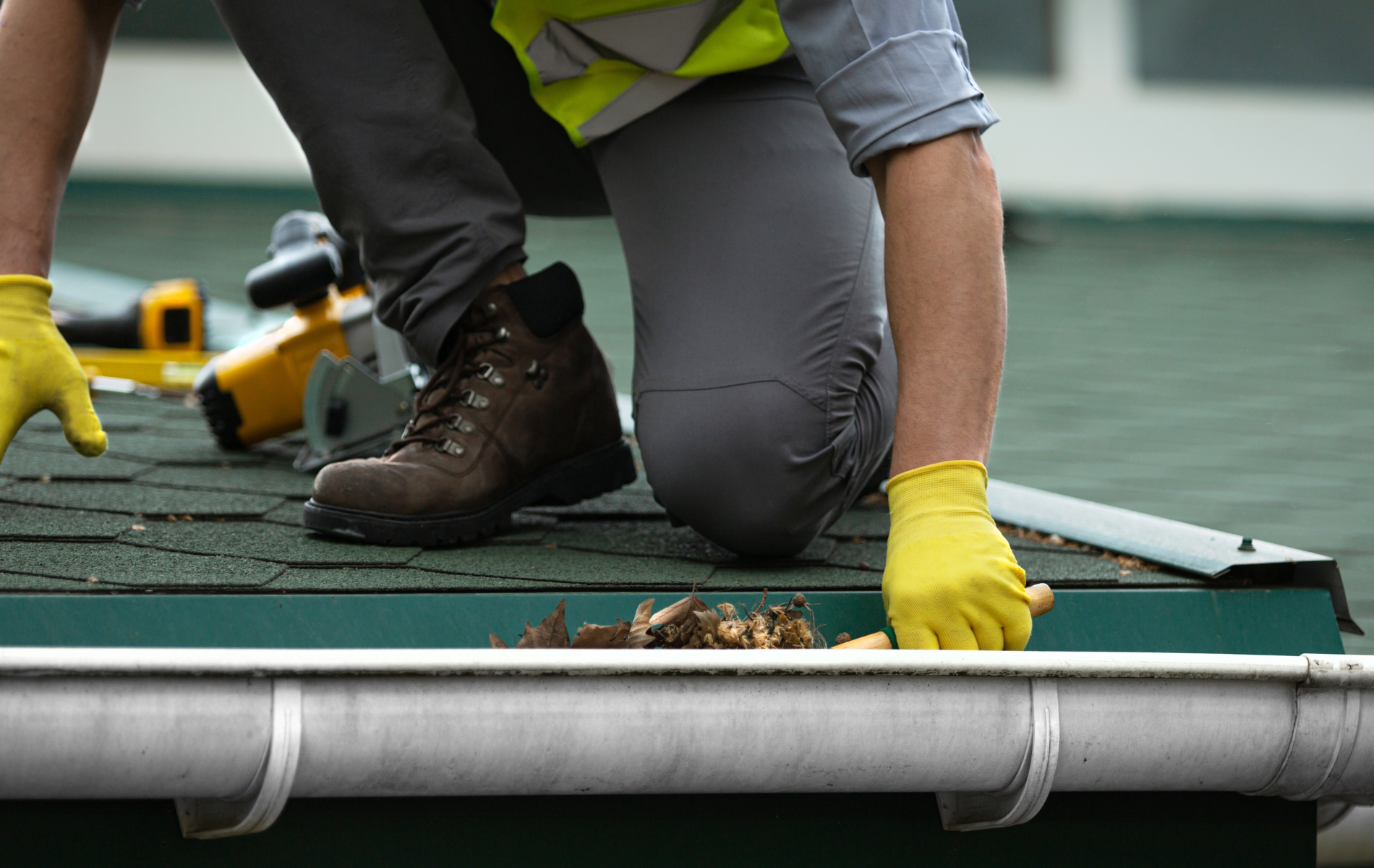 Person kneeling on a roof cleaning a gutter, wearing safety gear.