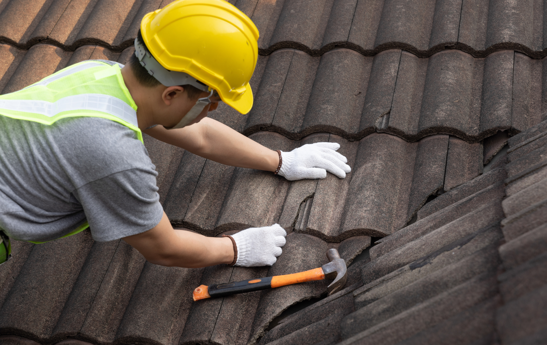 Roofer in yellow hard hat and gloves repairing a damaged tile roof with a hammer.