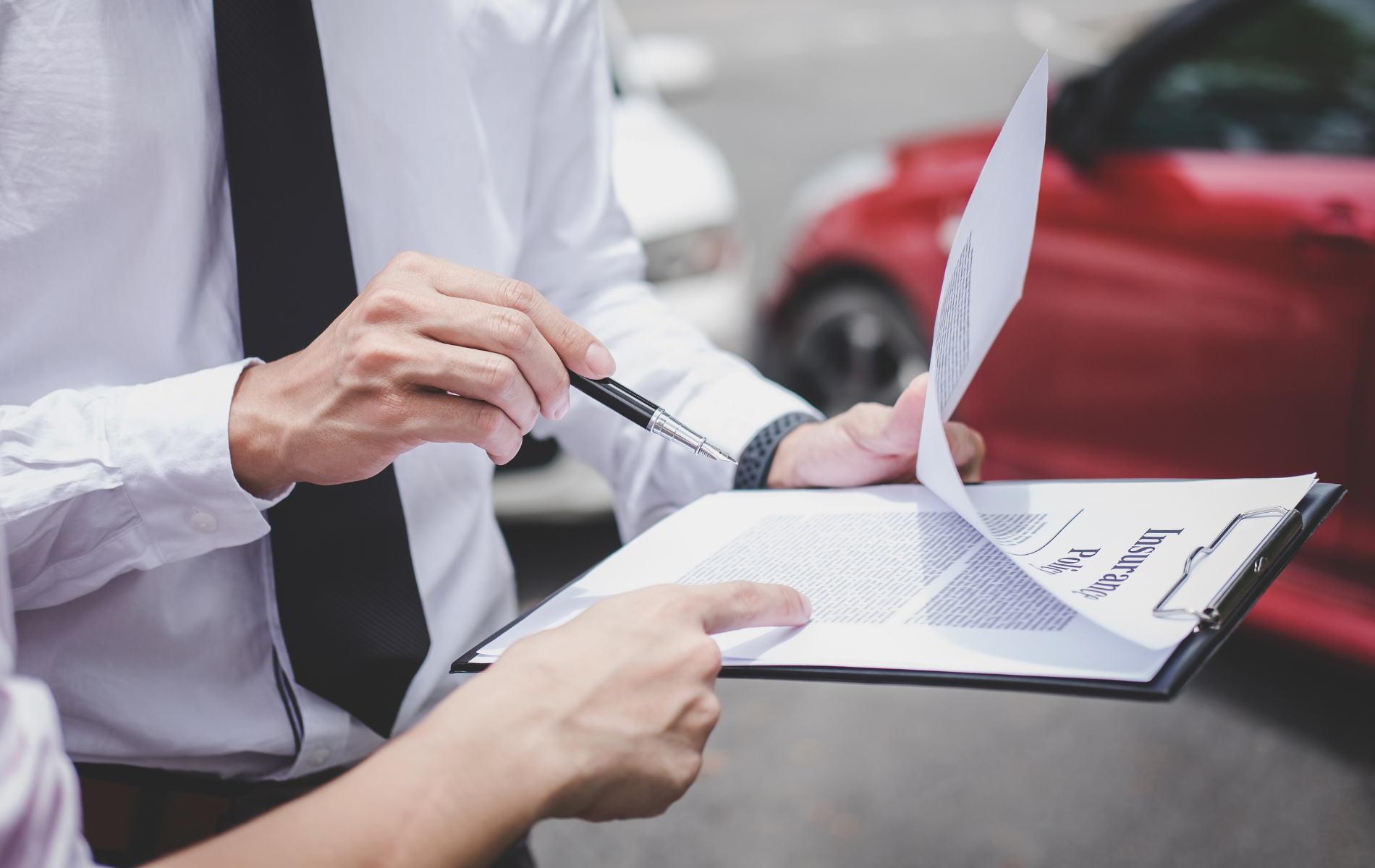 Two people reviewing paperwork at a car accident scene. One points with a pen near a red car.