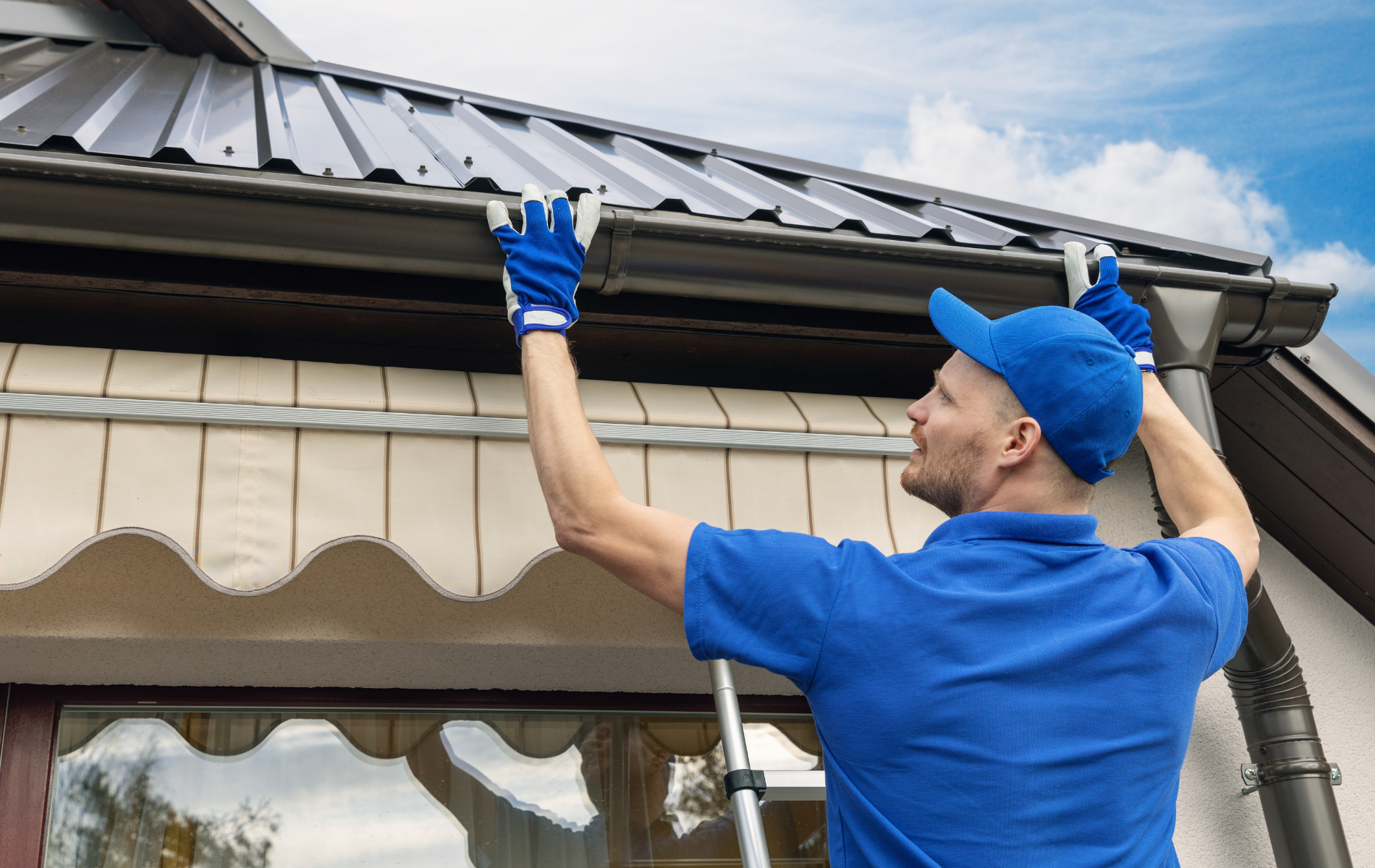 Man on ladder cleaning gutters, wearing blue shirt, gloves, and cap.