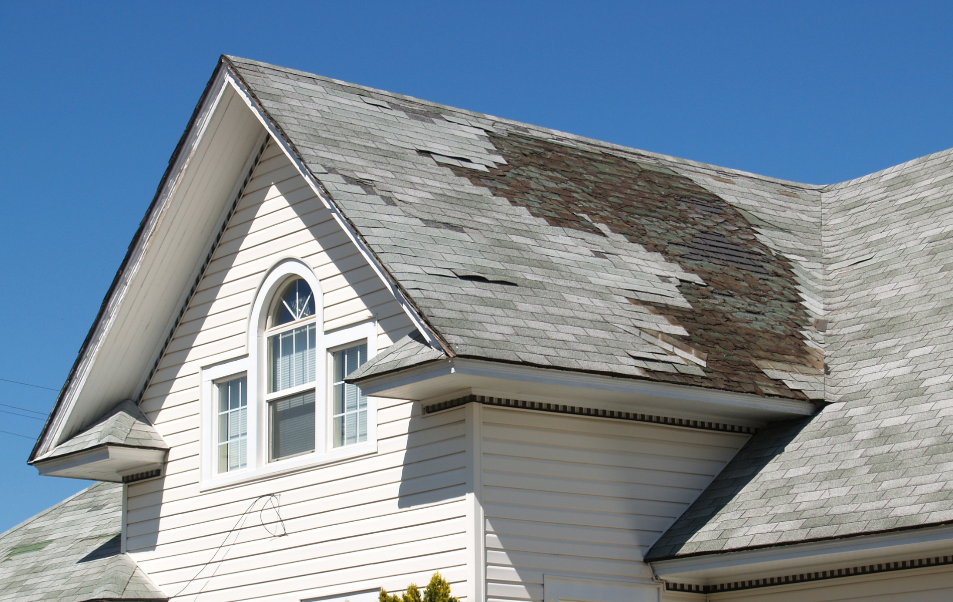 White house with severely damaged roof; dark patches, peeling shingles.