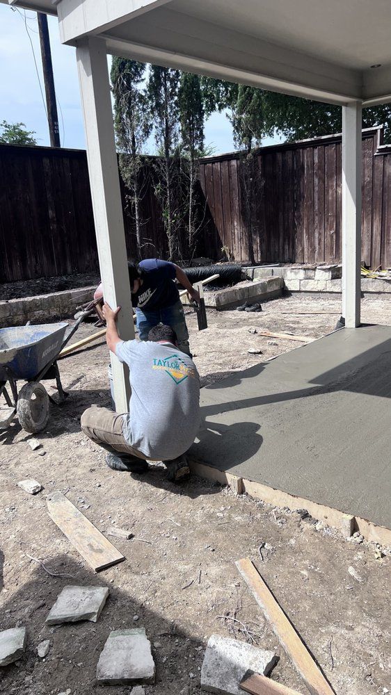 Two workers pouring concrete on a patio. Gray concrete, dirt ground, white posts, wooden fence.