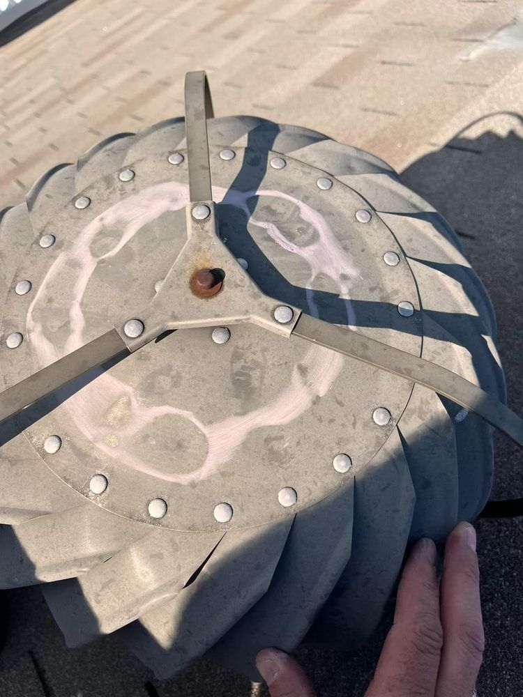 Close-up of a spinning roof vent on a weathered roof. White and pink stains on grey metal. Hand at the bottom right.