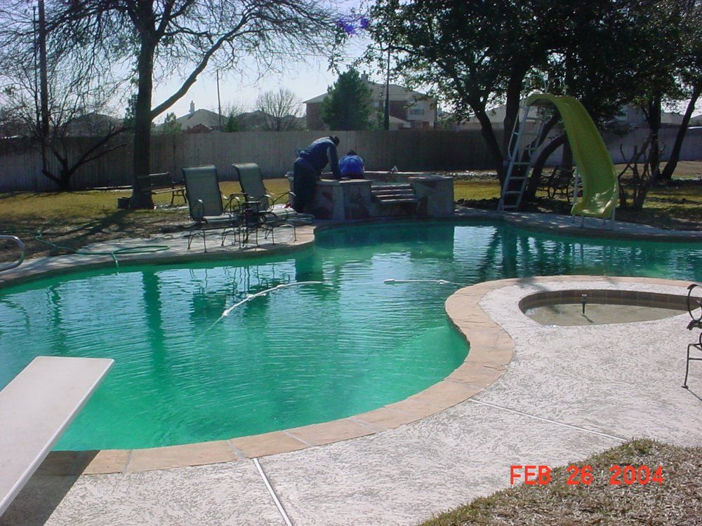 Swimming pool with a diving board, slide, and people near a small waterfall feature.