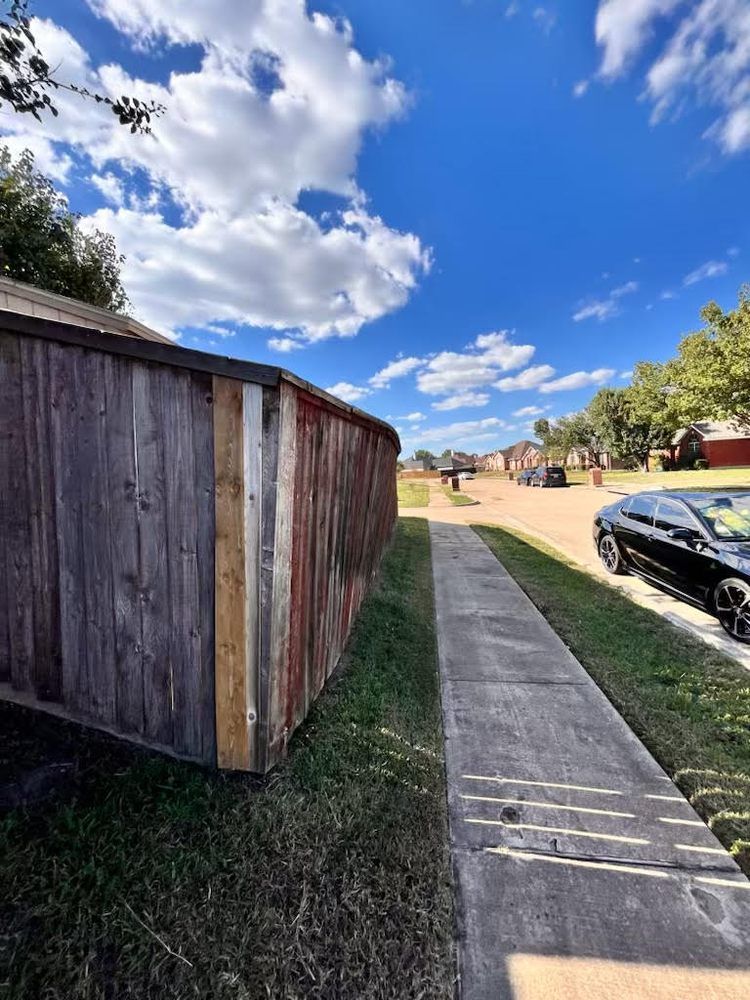 Weathered wooden fence next to a sidewalk, street with houses, blue sky with clouds.