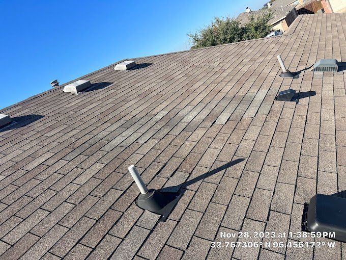 Brown asphalt shingle roof with vents and pipes under a clear, blue sky.